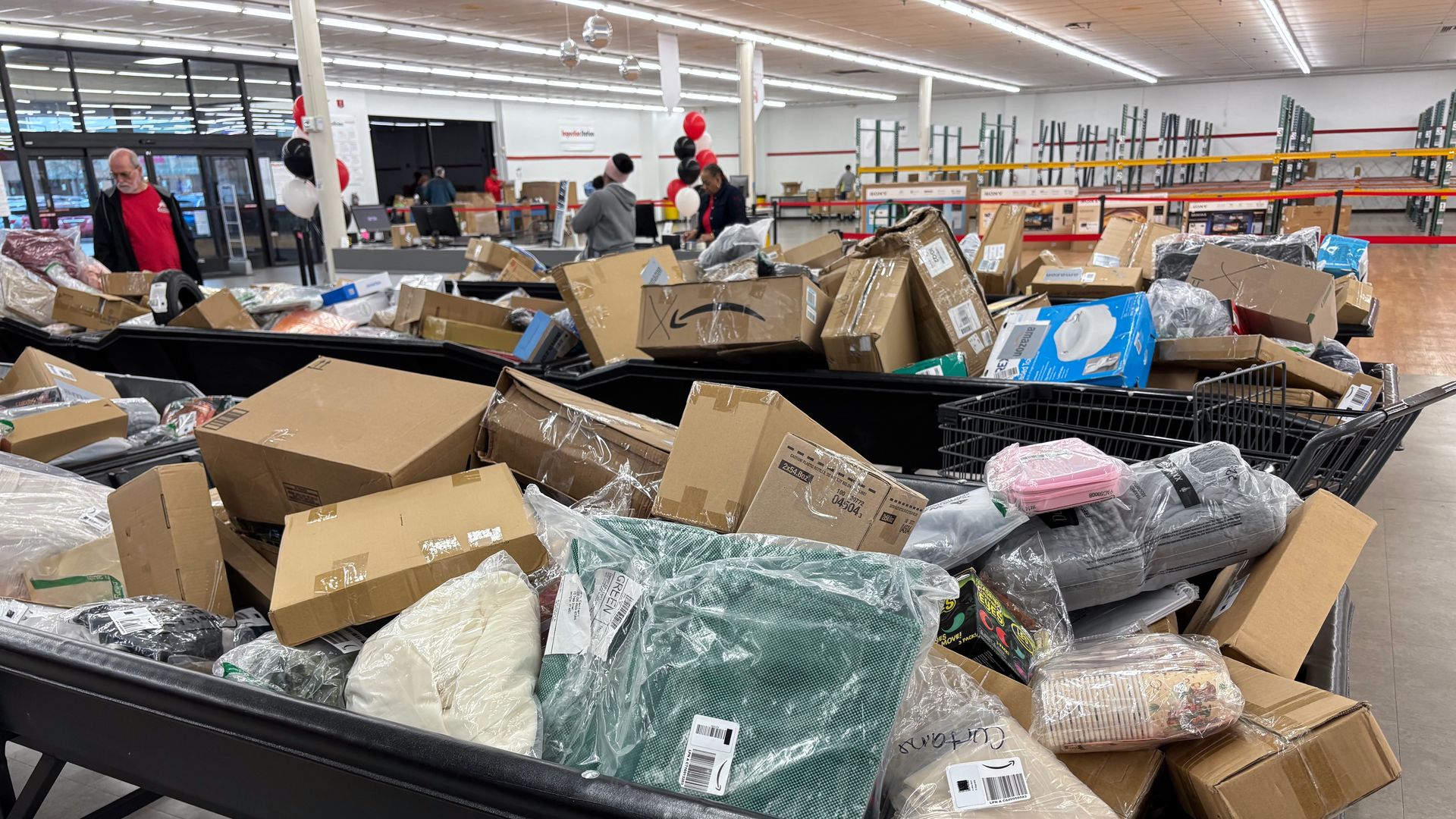 Large black bins filled with numerous cardboard boxes and plastic-wrapped items in a well-lit warehouse or store setting, with people and balloons visible in the background.