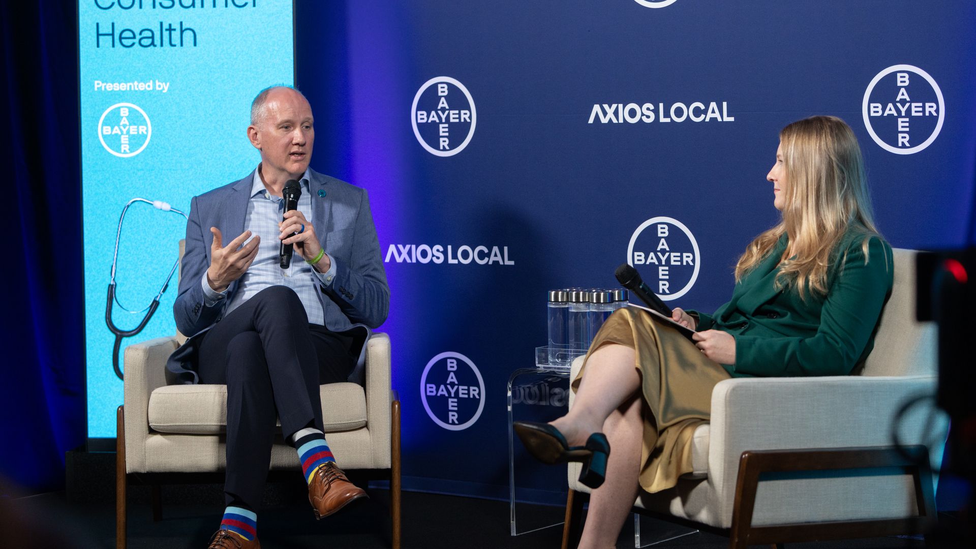 Two panelists on a stage with a blue backdrop showing "AXIOS LOCAL" and Bayer logos; a man in a blue blazer speaks into a microphone, while a woman in green listens.