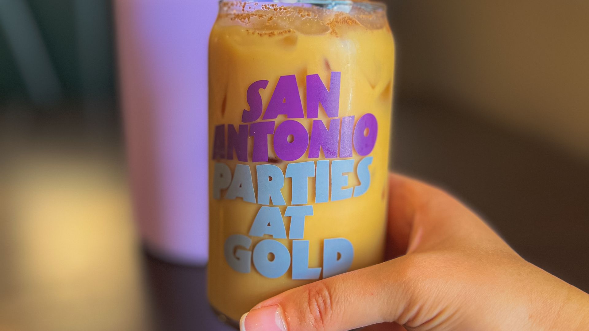 A hand holding a glass cup filled with coffee that says "San Antonio Parties at Gold" in purple font. 