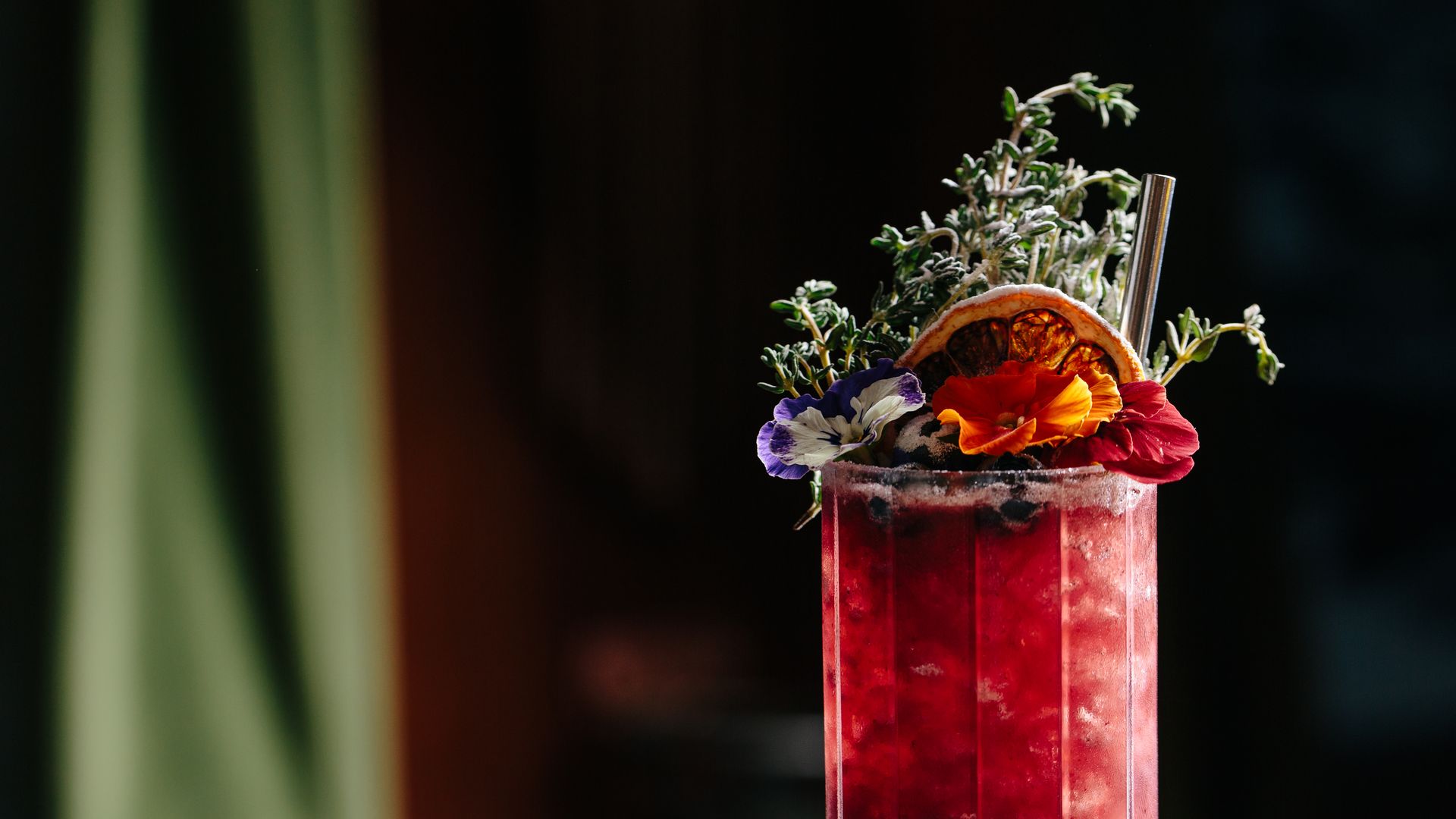 A tall glass with a red cocktail topped with fruit, flowers and herbs as garnish. 