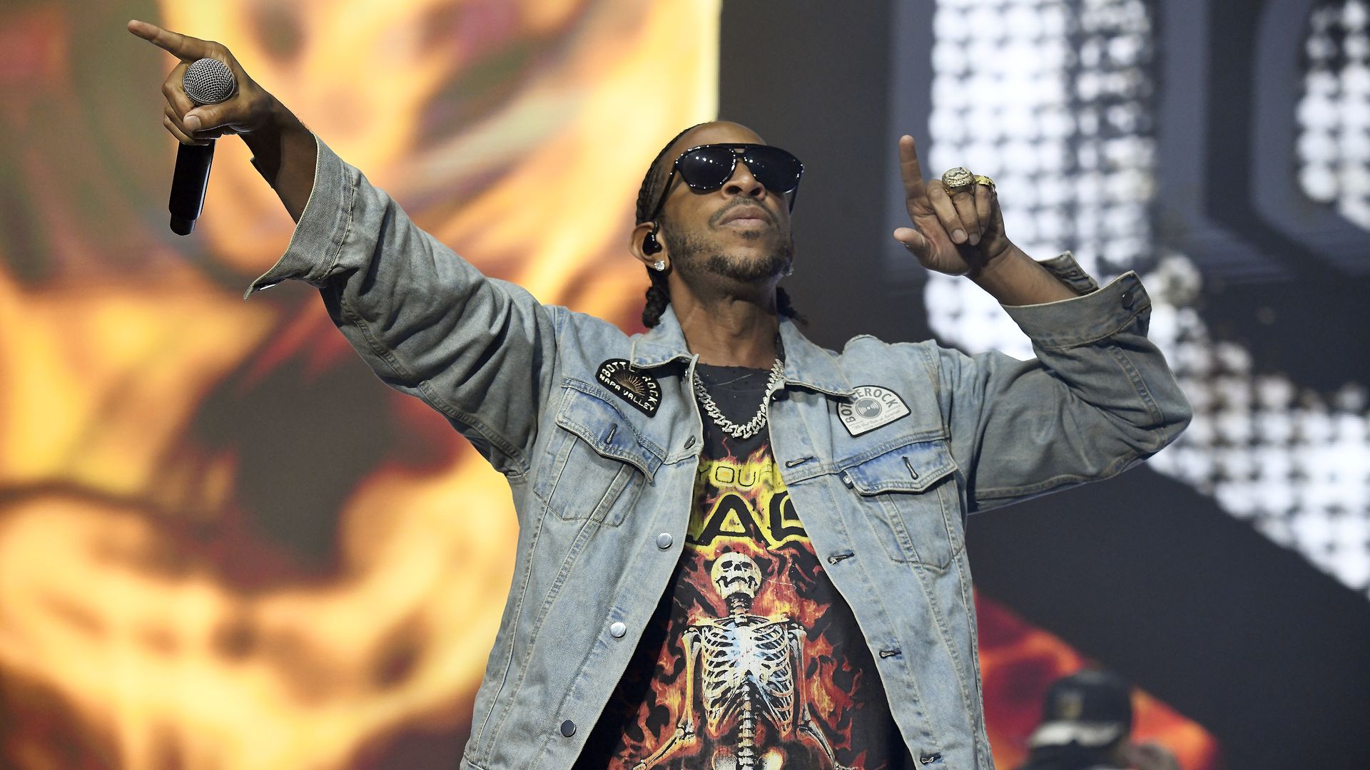A man wearing sunglasses, a blue jean jacket, necklace and a black, red and yellow T-shirt with a skull on the front holds a microphone in his right hand and points to the concert audience with his left hand.
