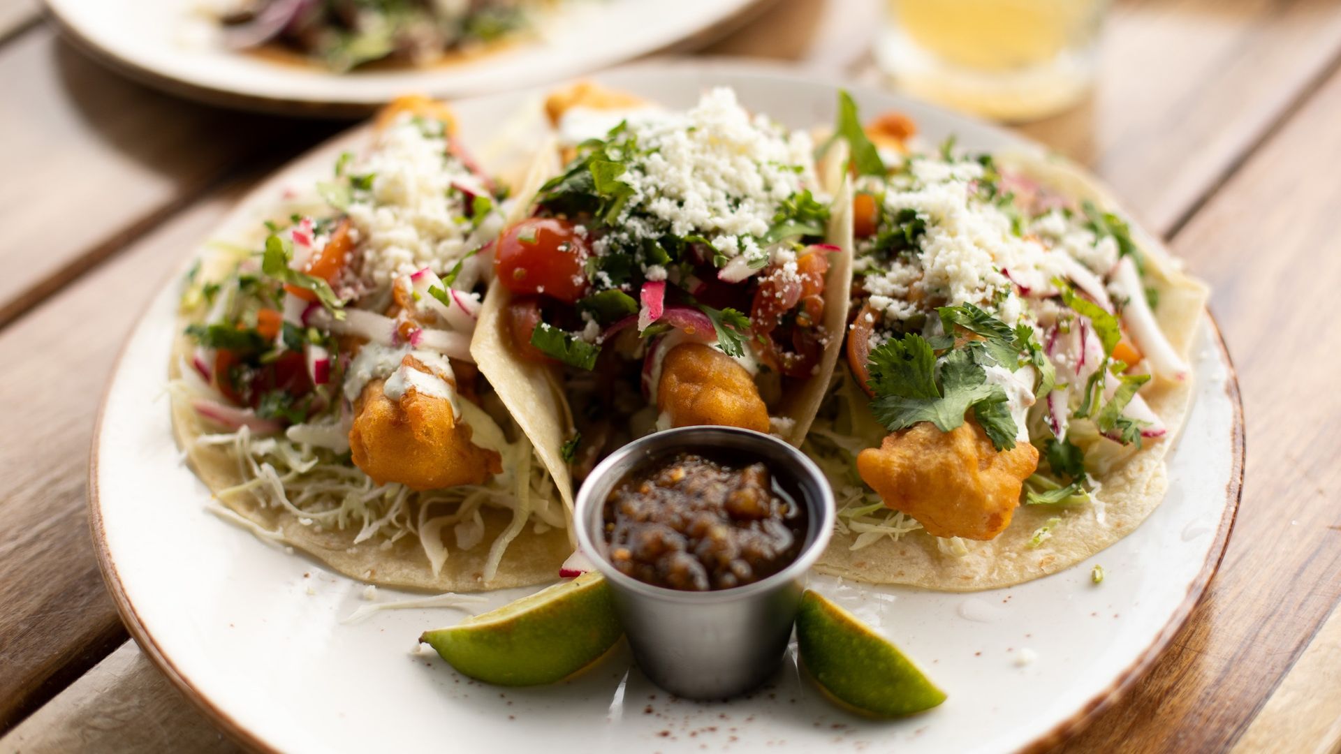 Three fried fish tacos in flour torillas topped with salsa, cabbage slaw and cotija cheese sit on a plate with a side of salsa and limes. 