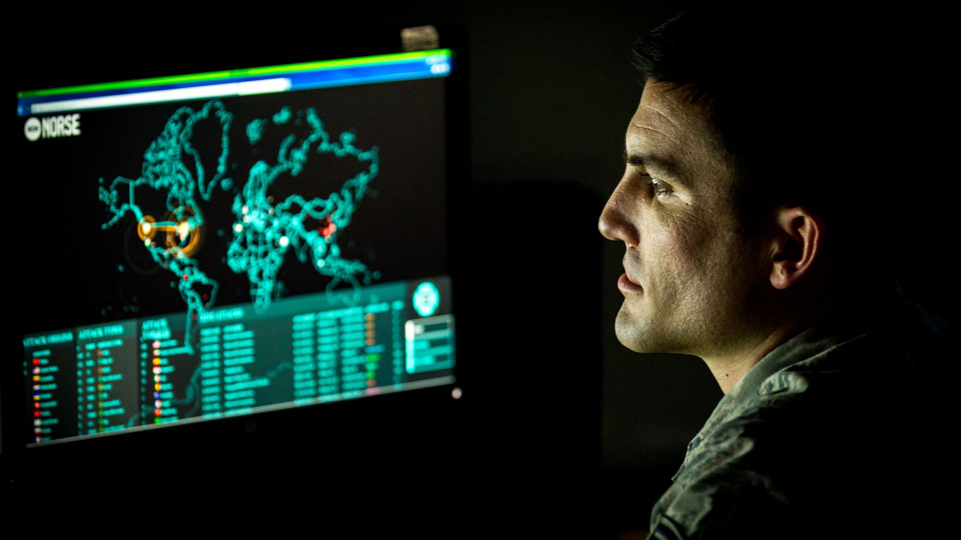 A computer screen displaying cyberattacks is seen at left. A man's face is illuminated by the screen at right. The background is pitch black.