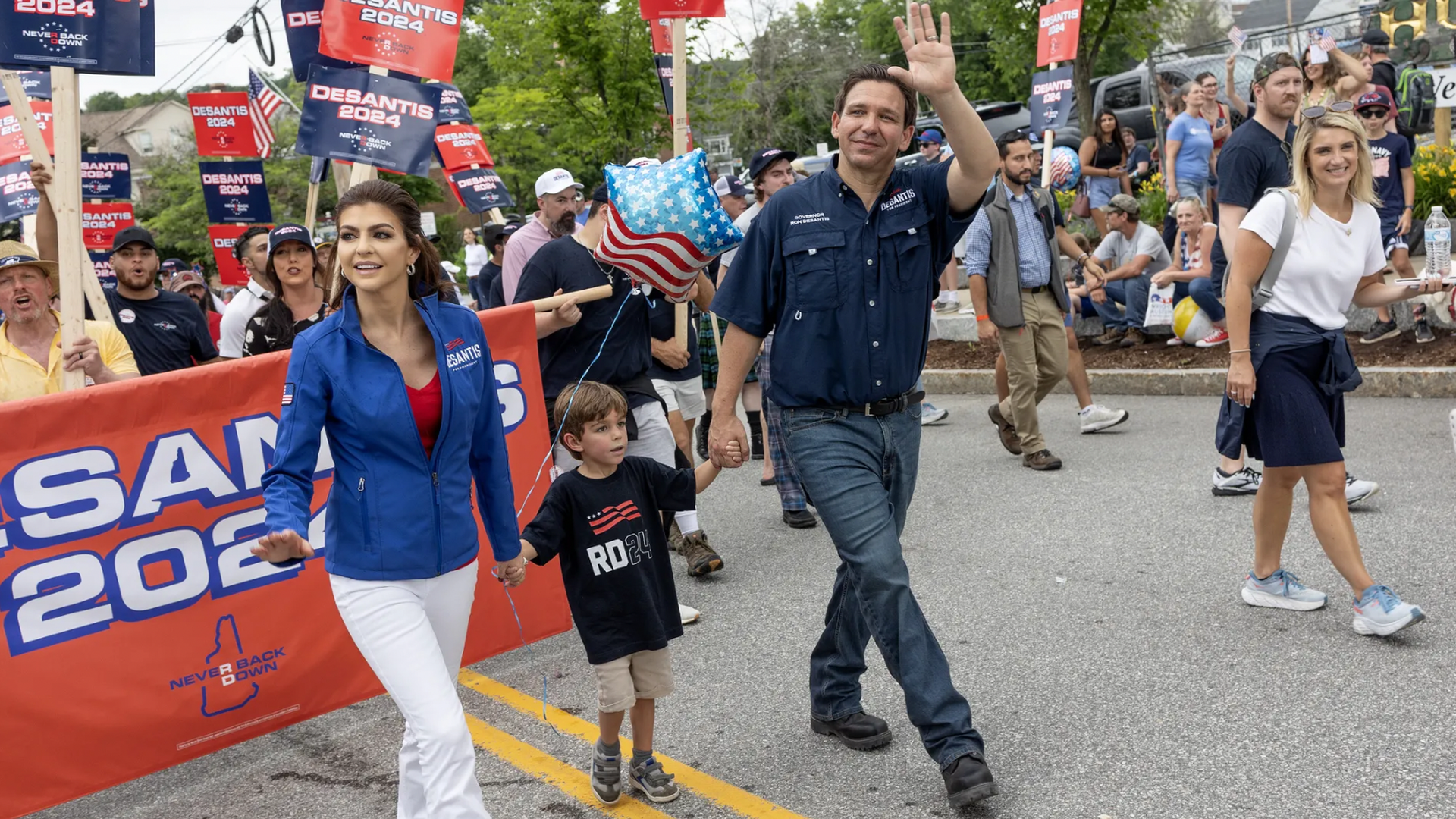 Ron Desantis walks in a parade with wife wearing blue jacket and son