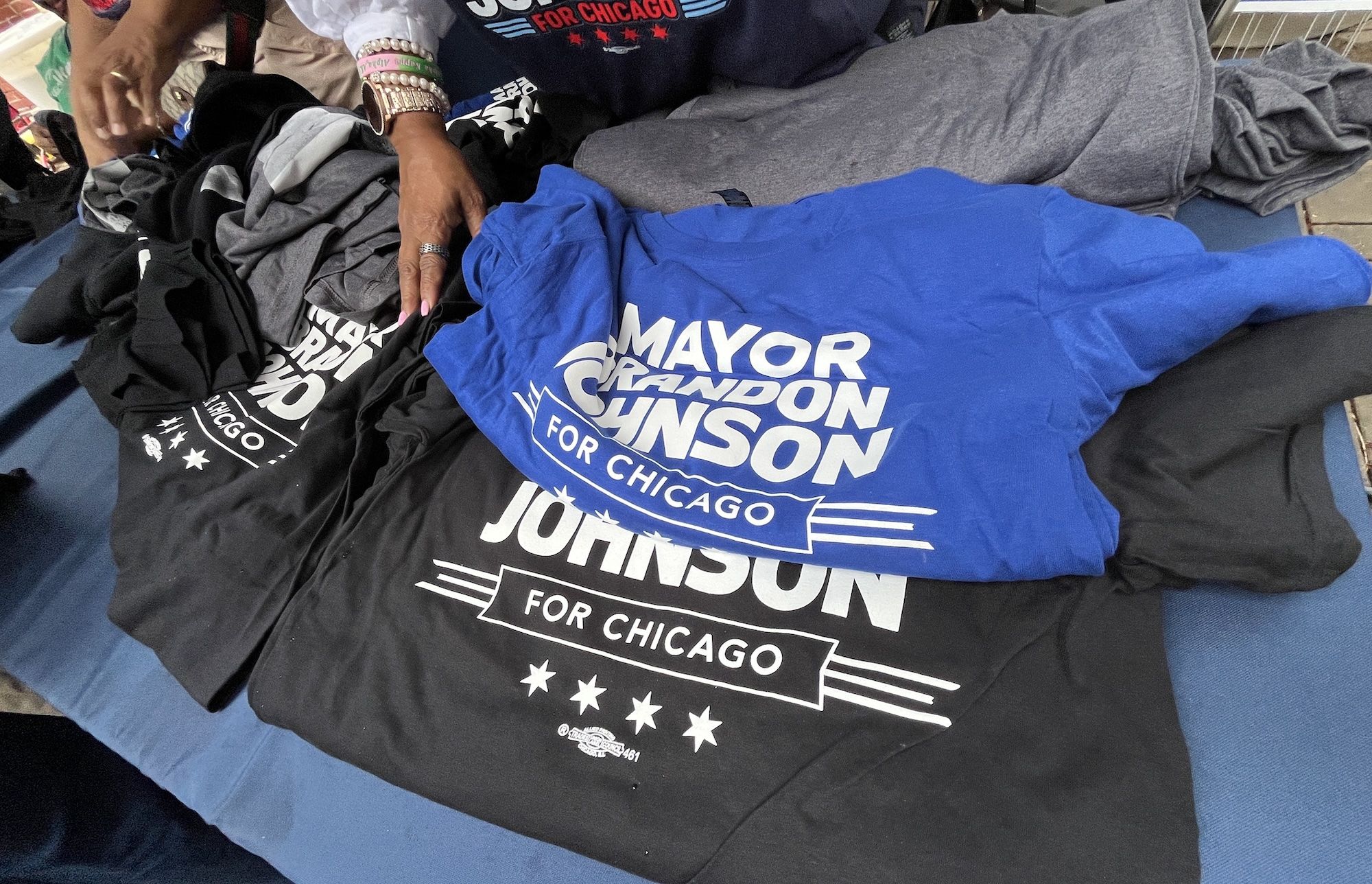 Pile of folded t-shirts on a blue surface; a royal blue shirt in the foreground reads "MAYOR JOHNSON FOR CHICAGO" in white letters. A hand with bracelets rests nearby.