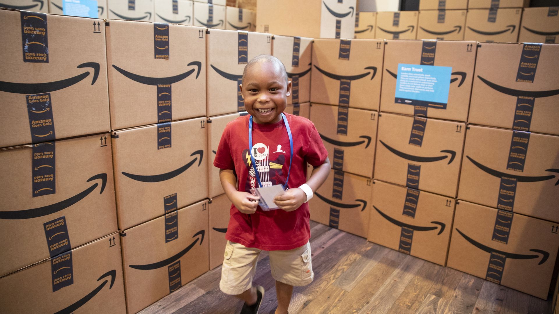 A boy in a red shirt stands in front a pile of Amazon boxes