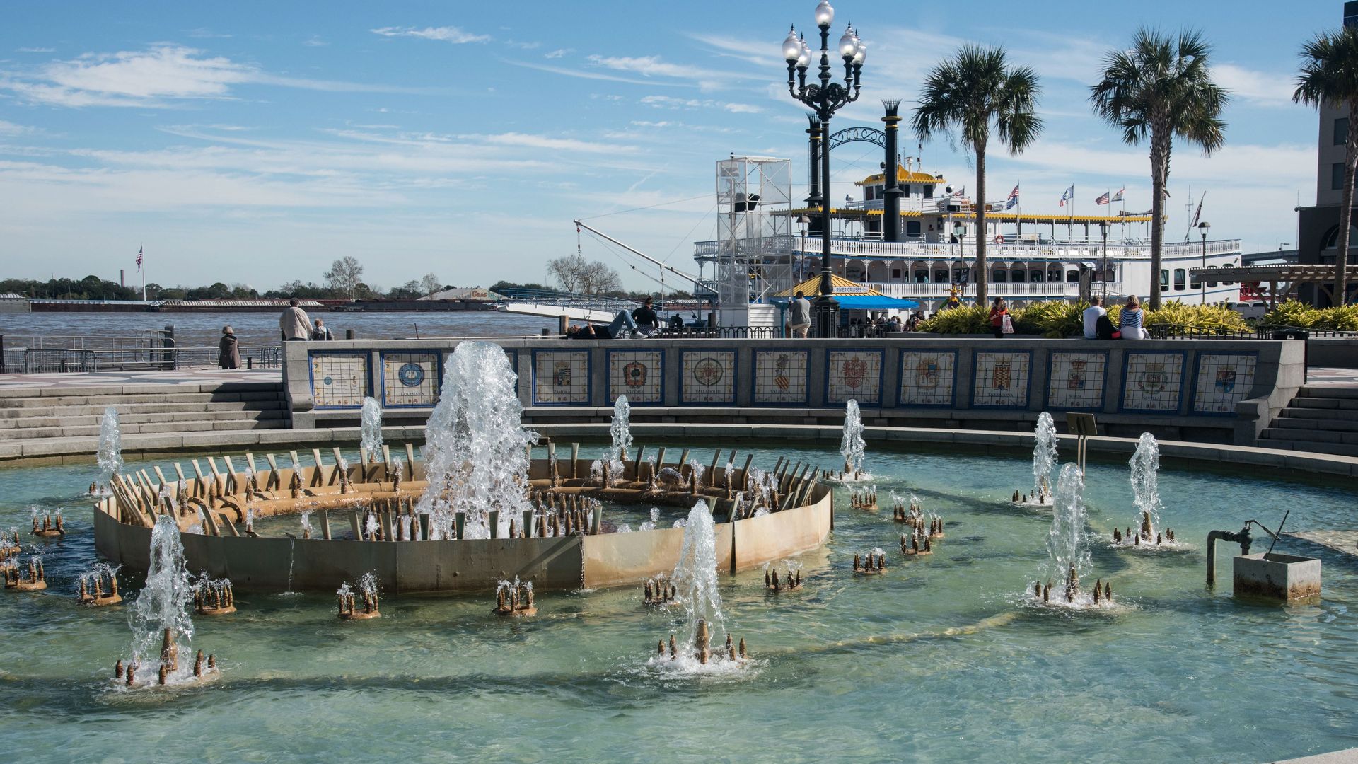 Photo shows a large fountain in the center of Spanish Plaza. A paddlewheel in in the Mississippi River in the background.