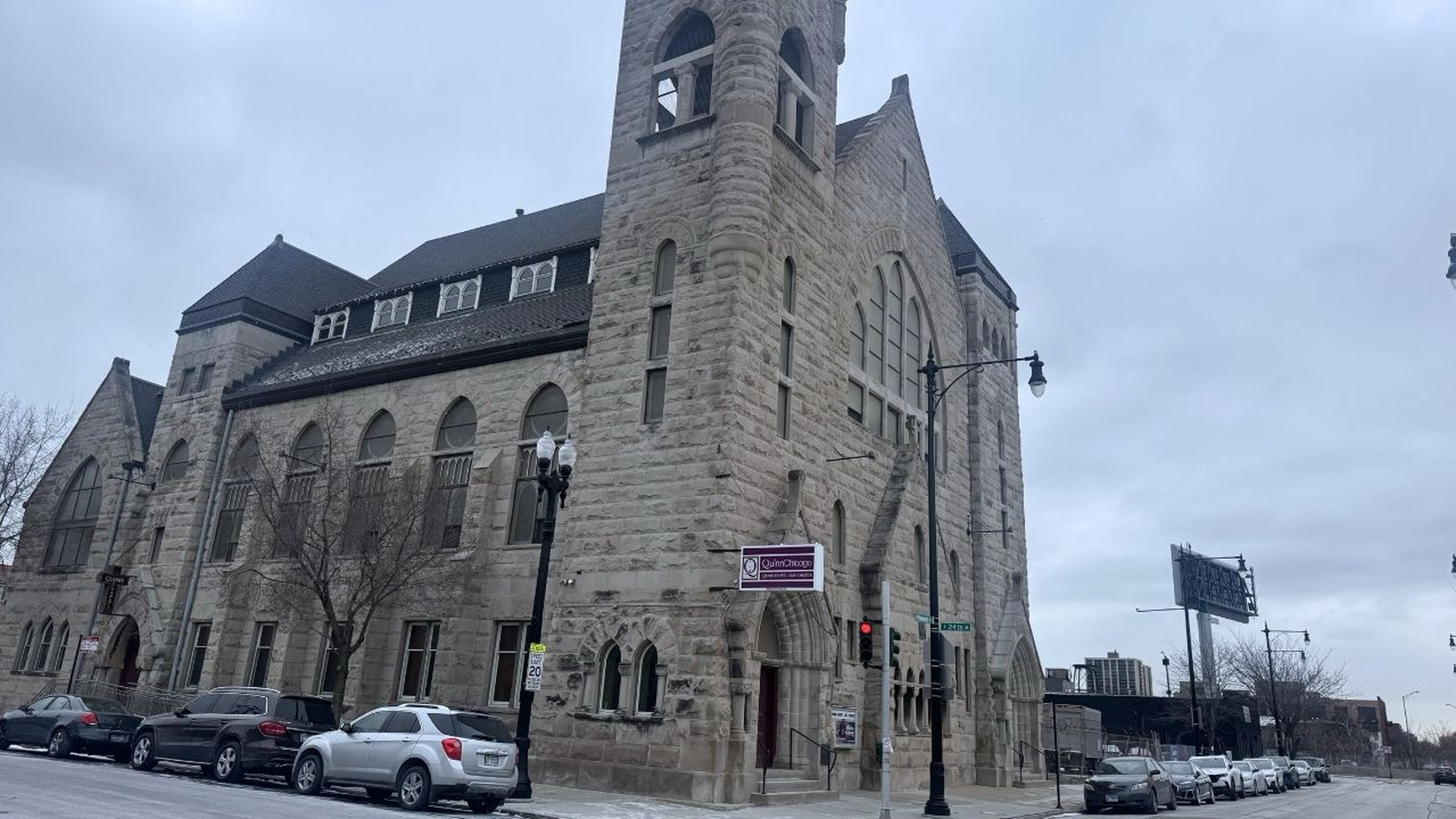Large gray stone building with Gothic-style architecture, arched windows, and a tower on the corner; cars parked along the street; cloudy sky overhead.