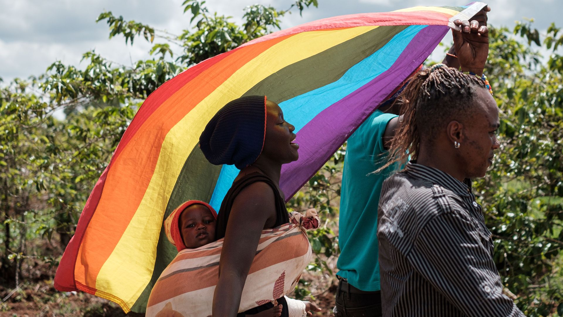 In this image, a man carries a rainbow flag while a woman carrying a baby in a papoose follows behind him.