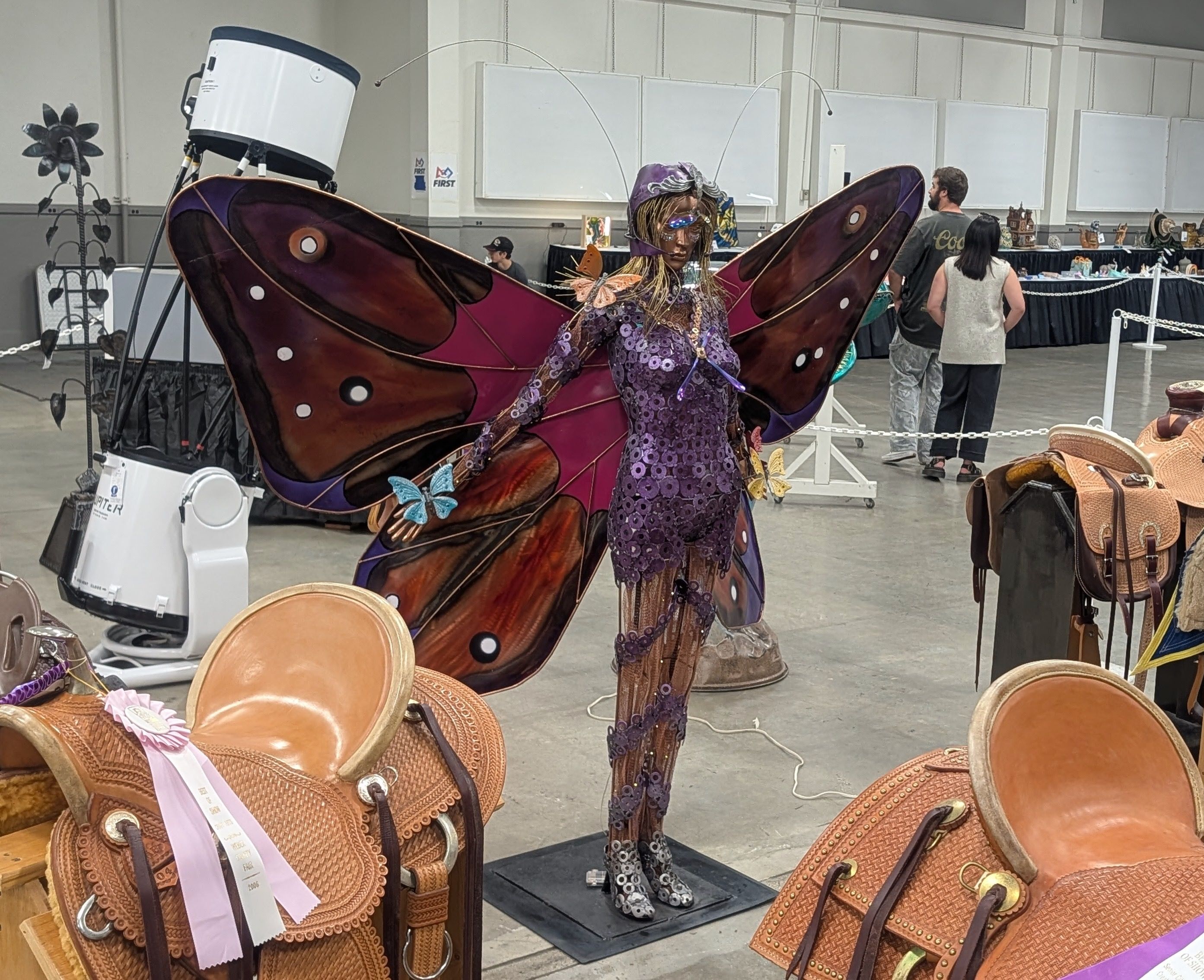 A person in a purple, sequined butterfly costume with large wings and a jeweled headpiece stands in a convention hall, surrounded by display booths and leather saddles.