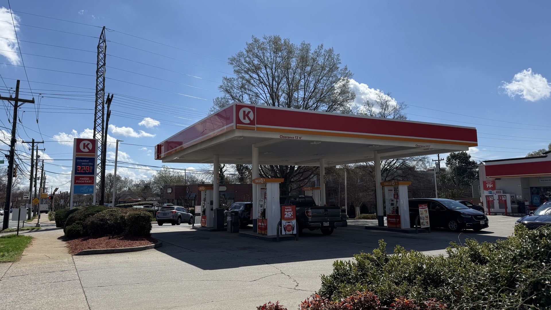 Bright day at a Circle K gas station with red and white canopy, several cars refueling, green bushes, and a price sign showing $3.29 for unleaded gas under a clear blue sky.
