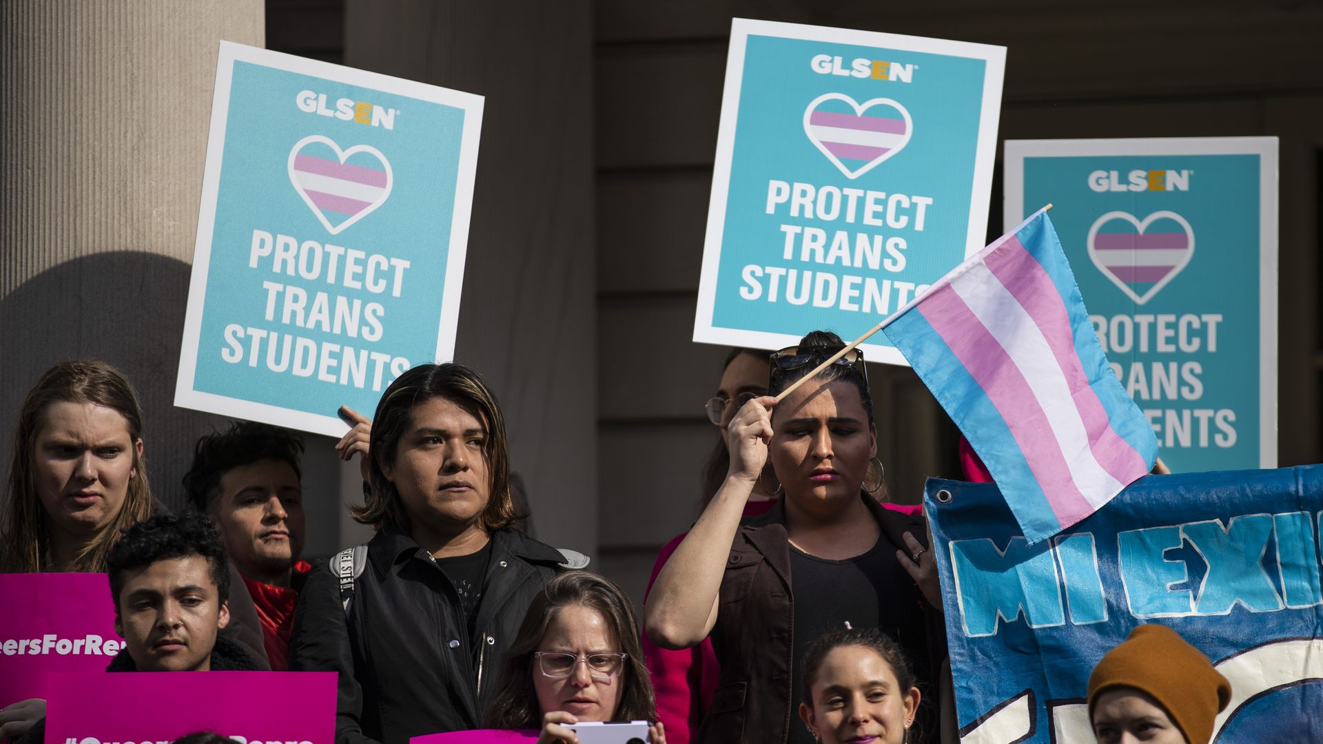 In this image, a transgender pride flag is waved while others hold signs that read "protect trans students"