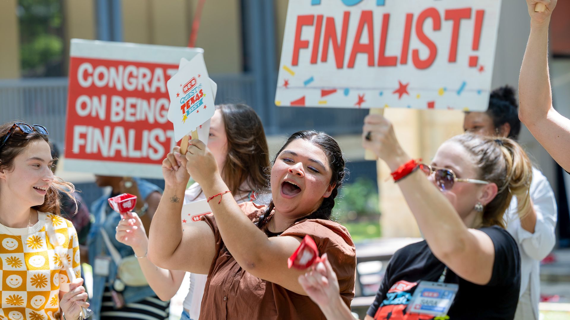 Joyful outdoor crowd of diverse people holding red-and-white signs reading "FINALIST!" and "CONGRATS ON BEING A FINALIST," with confetti and bright clothing.