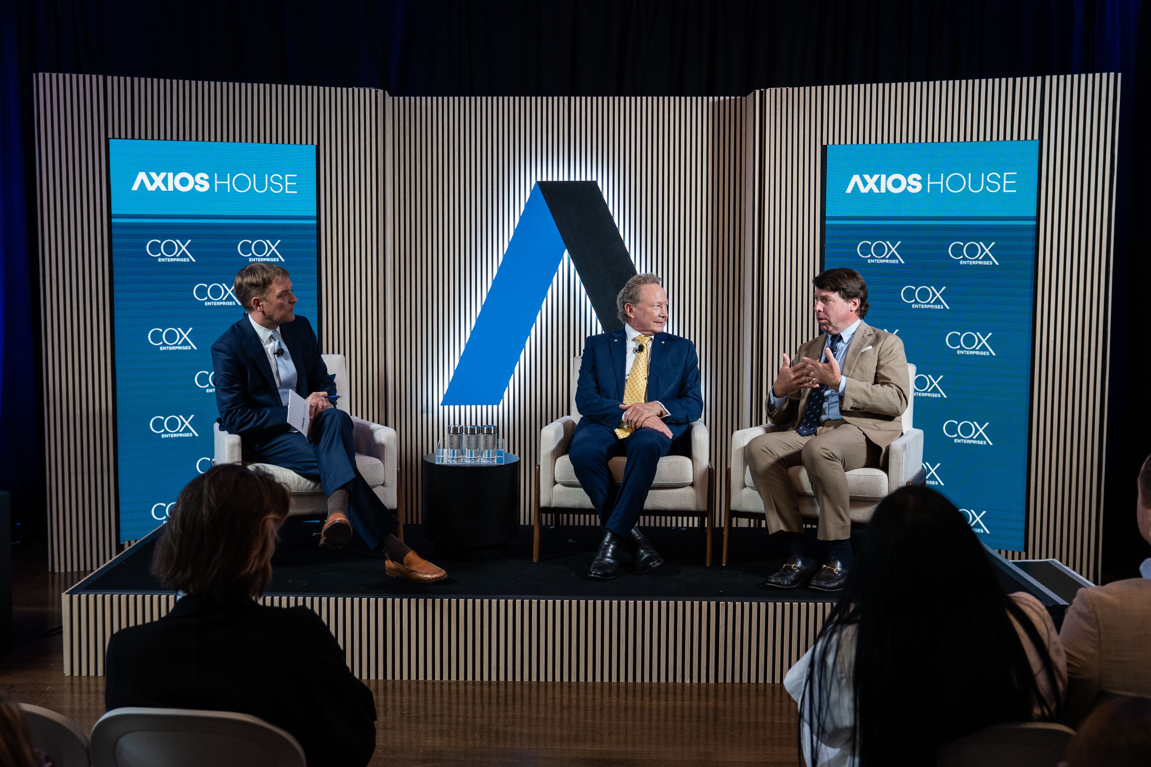 Panel of three men in suits seated on a stage with blue Axios House banners and Cox Enterprises logos; a blue and black diagonal sculpture sits between them, striped backdrop, audience visible.