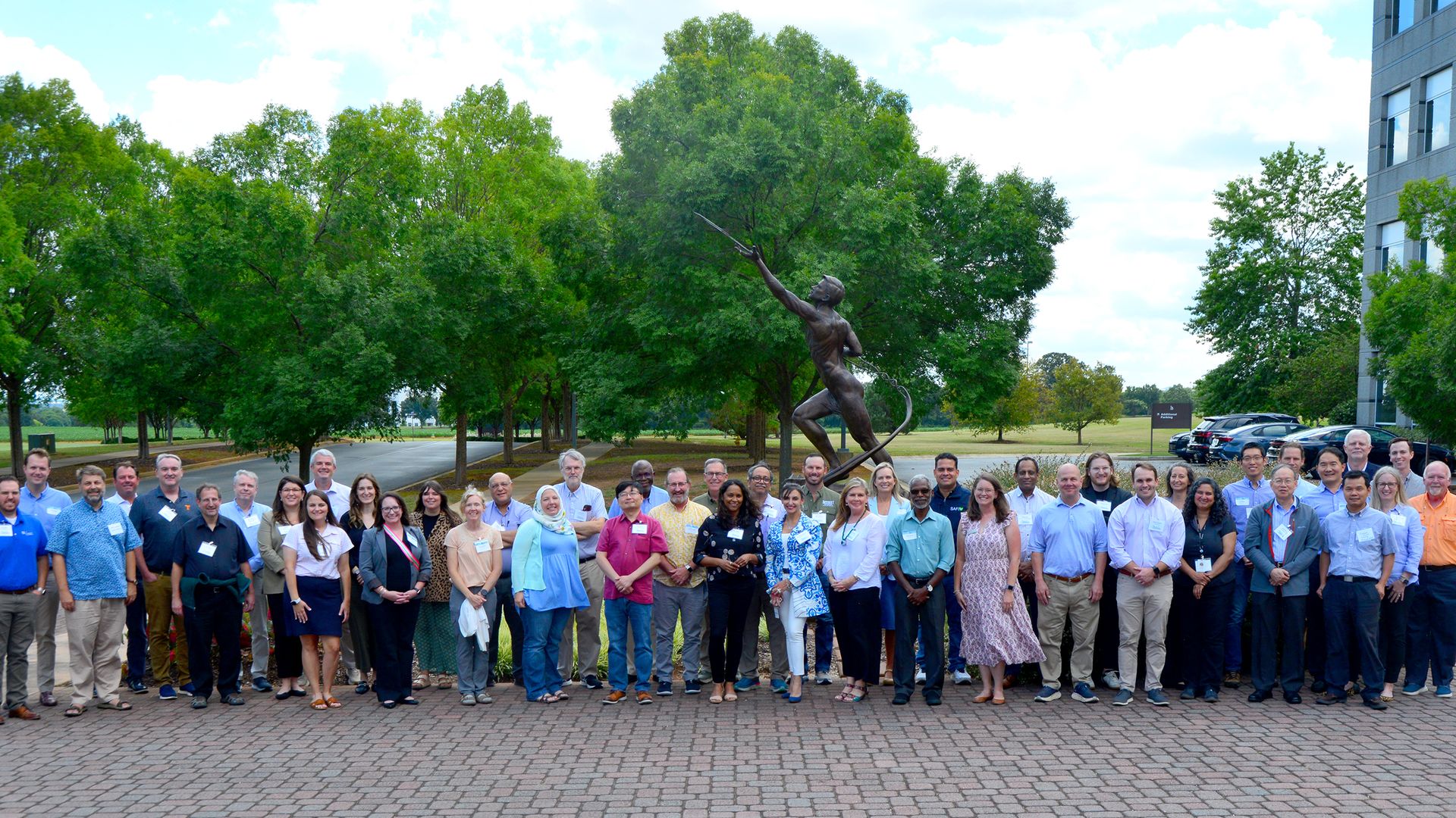 Large group of diverse adults posing outdoors on a paved area in front of a bronze statue of a man aiming a bow, with green trees and a building in the background under a partly cloudy sky.
