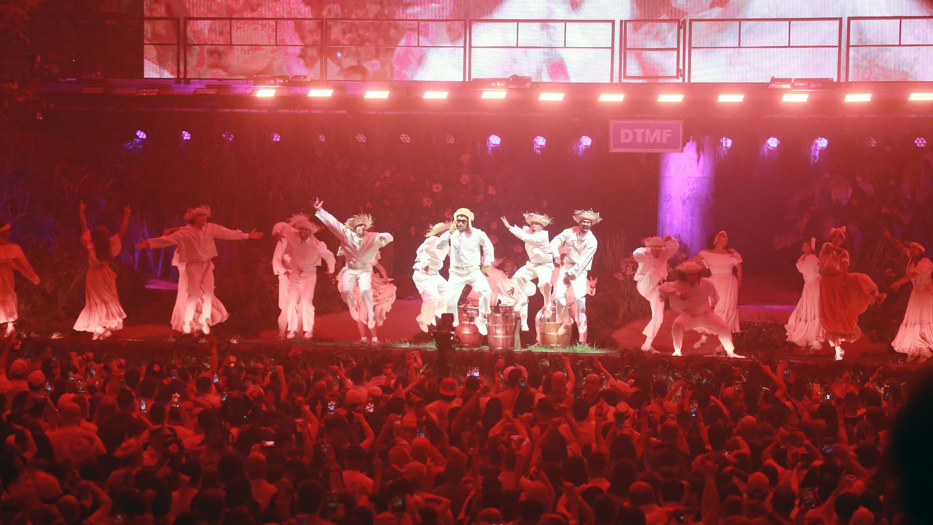 Performers wearing all white on a red-lit stage at a concert with hundreds of fans watching.