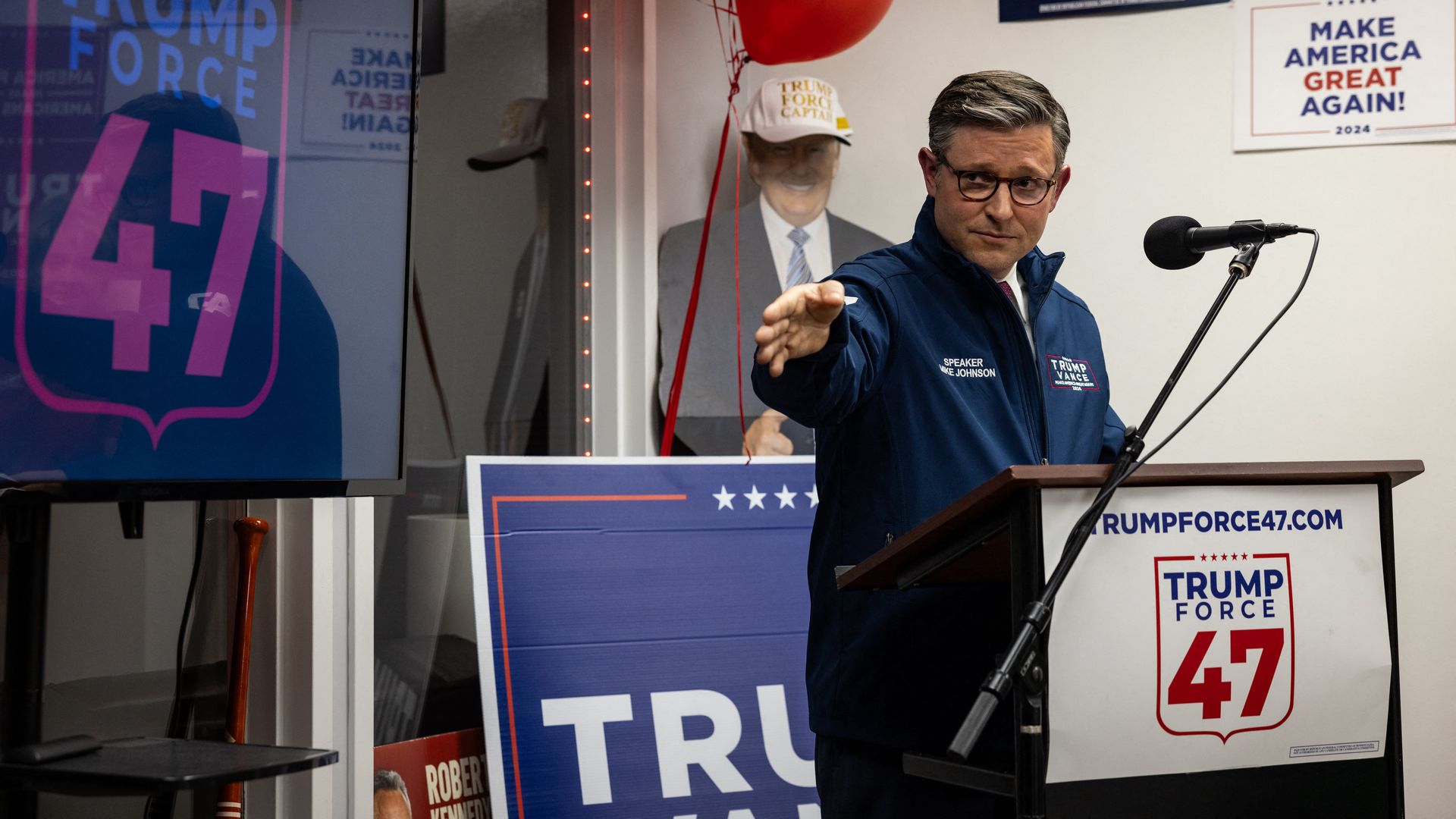 Mike Johnson, wearing a blue windbreaker and speaking at a "Trump Force 47" podium.