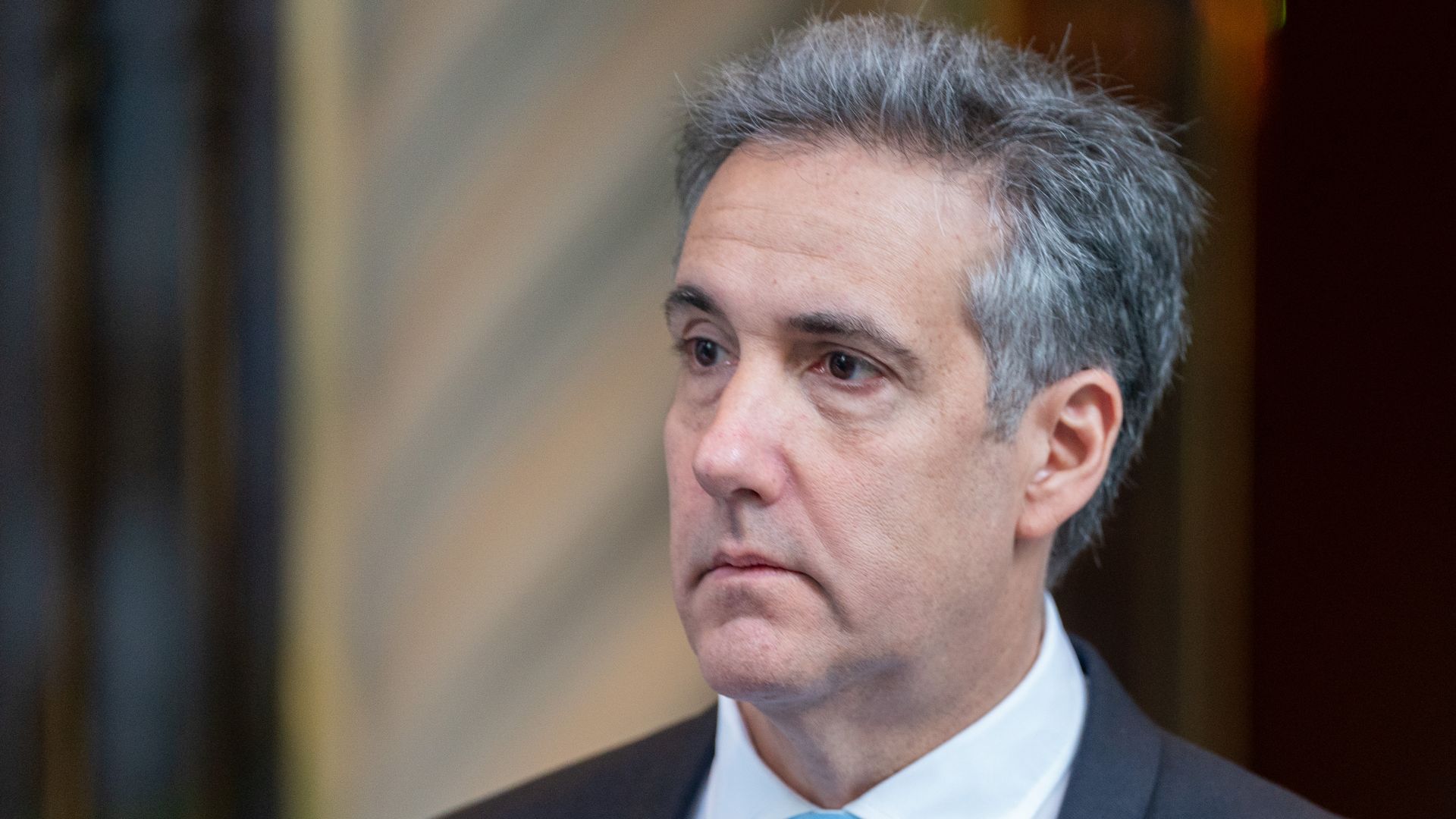 Close-up of a middle-aged man with gray hair, wearing a dark suit and light blue tie, looking slightly to the left. A blurred background behind him.