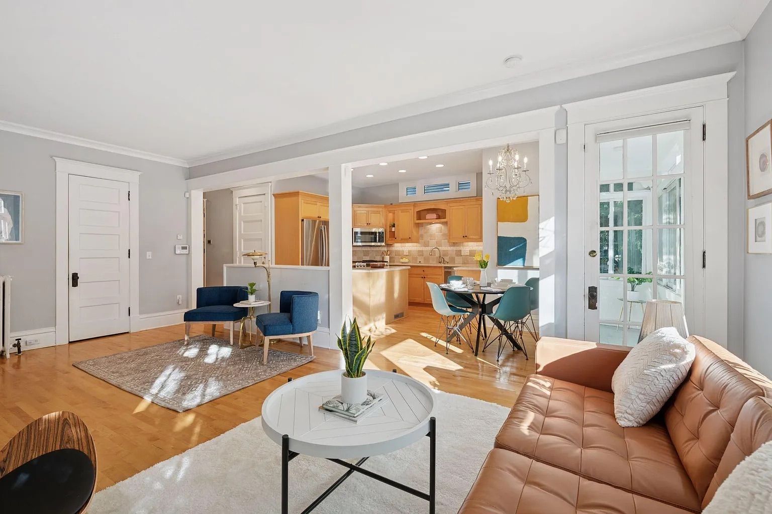 Bright open living room and kitchen with light gray walls, tan leather sofa, blue chairs, round white coffee table with plant, wooden floors, and glass door leading outside.