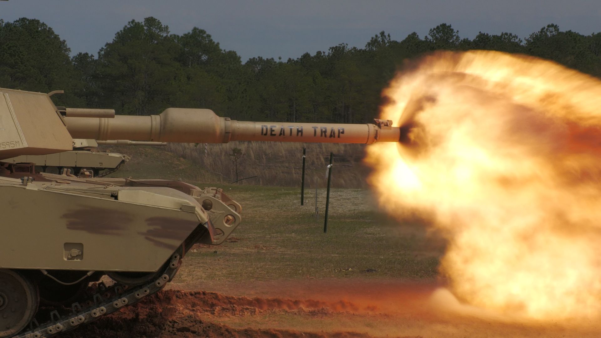 An Abrams tank takes a shot. Fire shoots out the barrel. "Death Trap" is written on the barrel, as well. A forest can be seen in the background.
