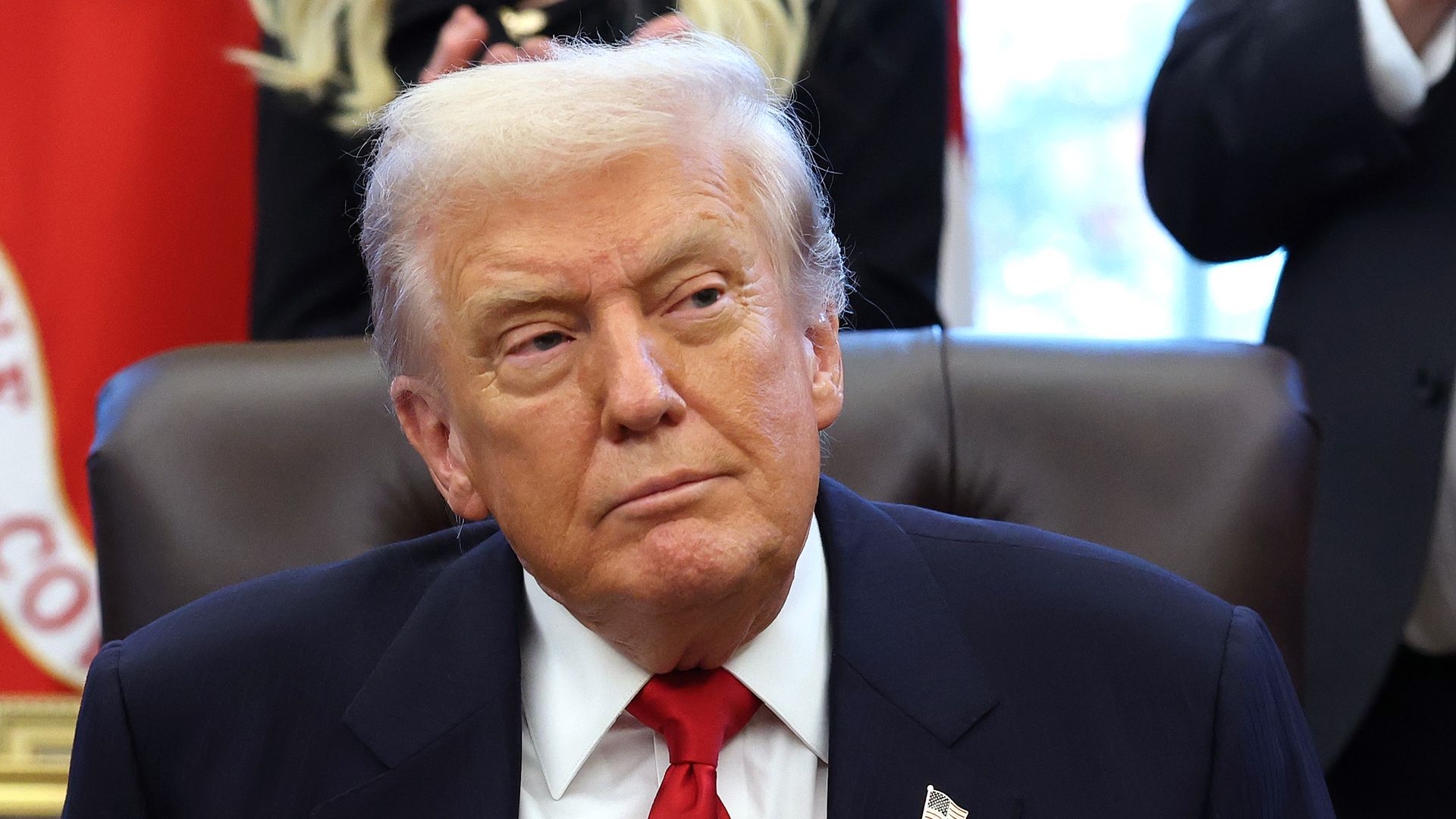 President Trump, wearing a navy jacket with a US flag pin at the top of his left lapel, white shirt and red tie, stares ahead from his brown leather chair in the White House Oval Office.