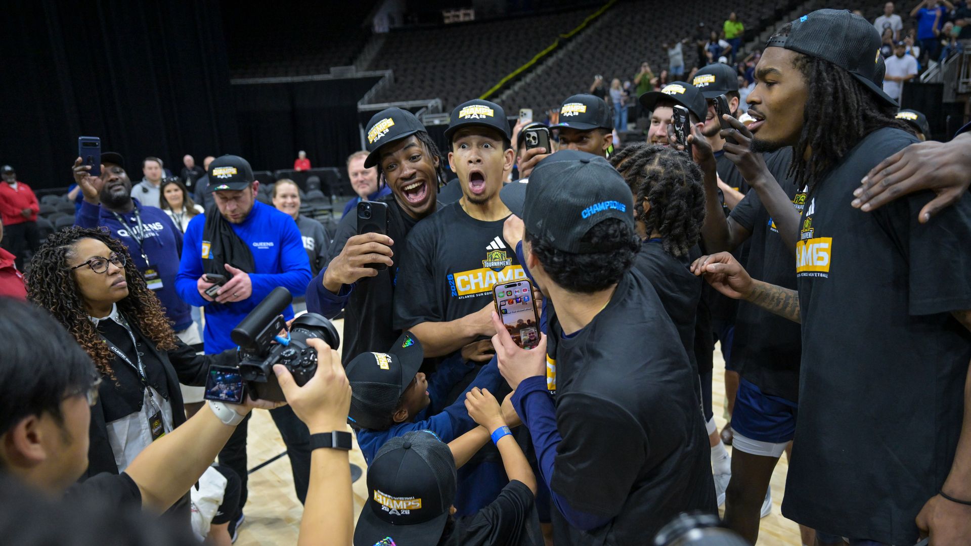 Group of basketball players in black shirts and caps celebrating on a court, wearing CHAMPS hats and taking selfies with phones and cameras while fans and photographers surround them.