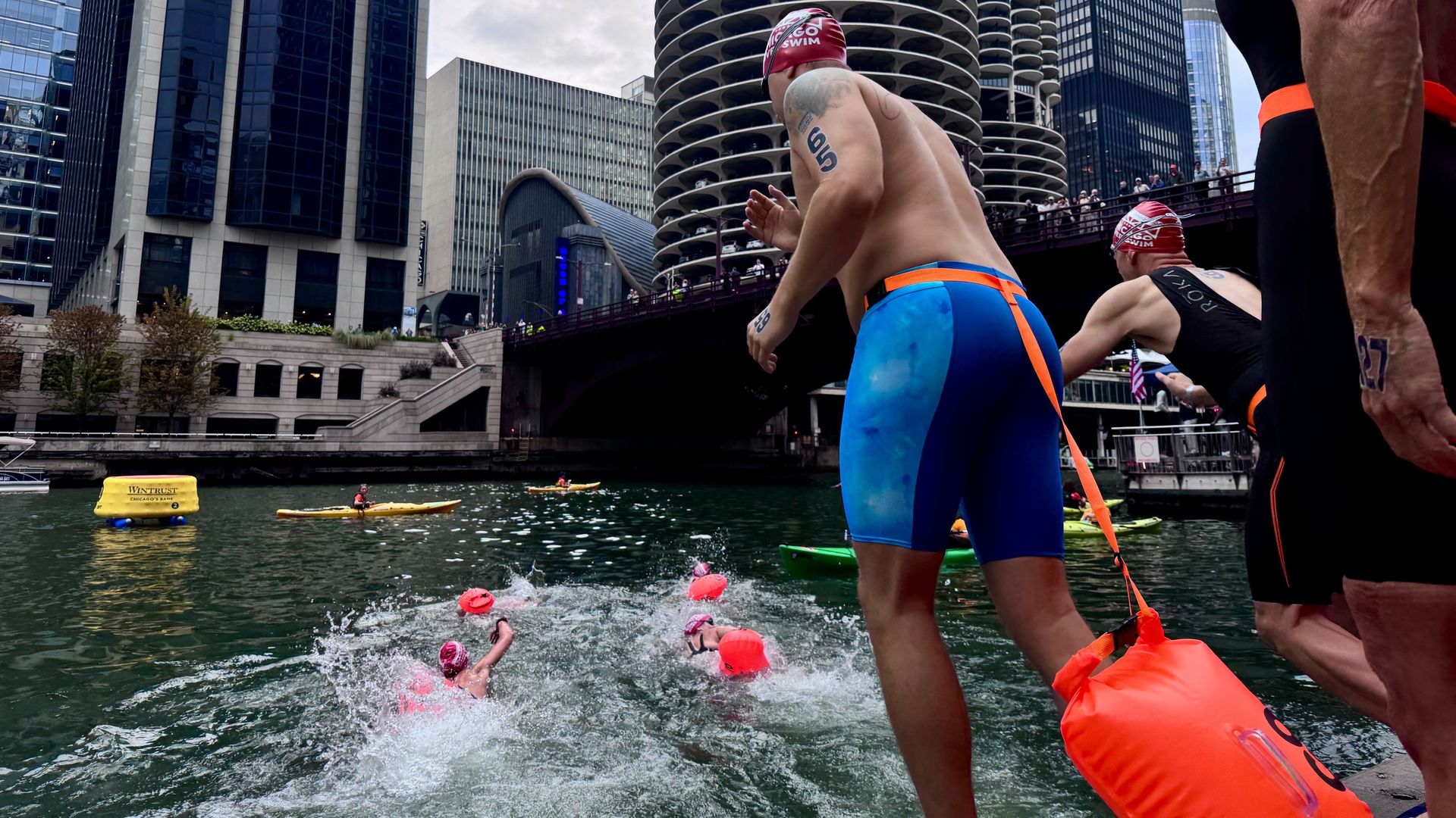 Swimmers in red caps and blue or black swimwear diving into a river in a city with tall buildings and unique round balconies, some spectators watching from a bridge above.
