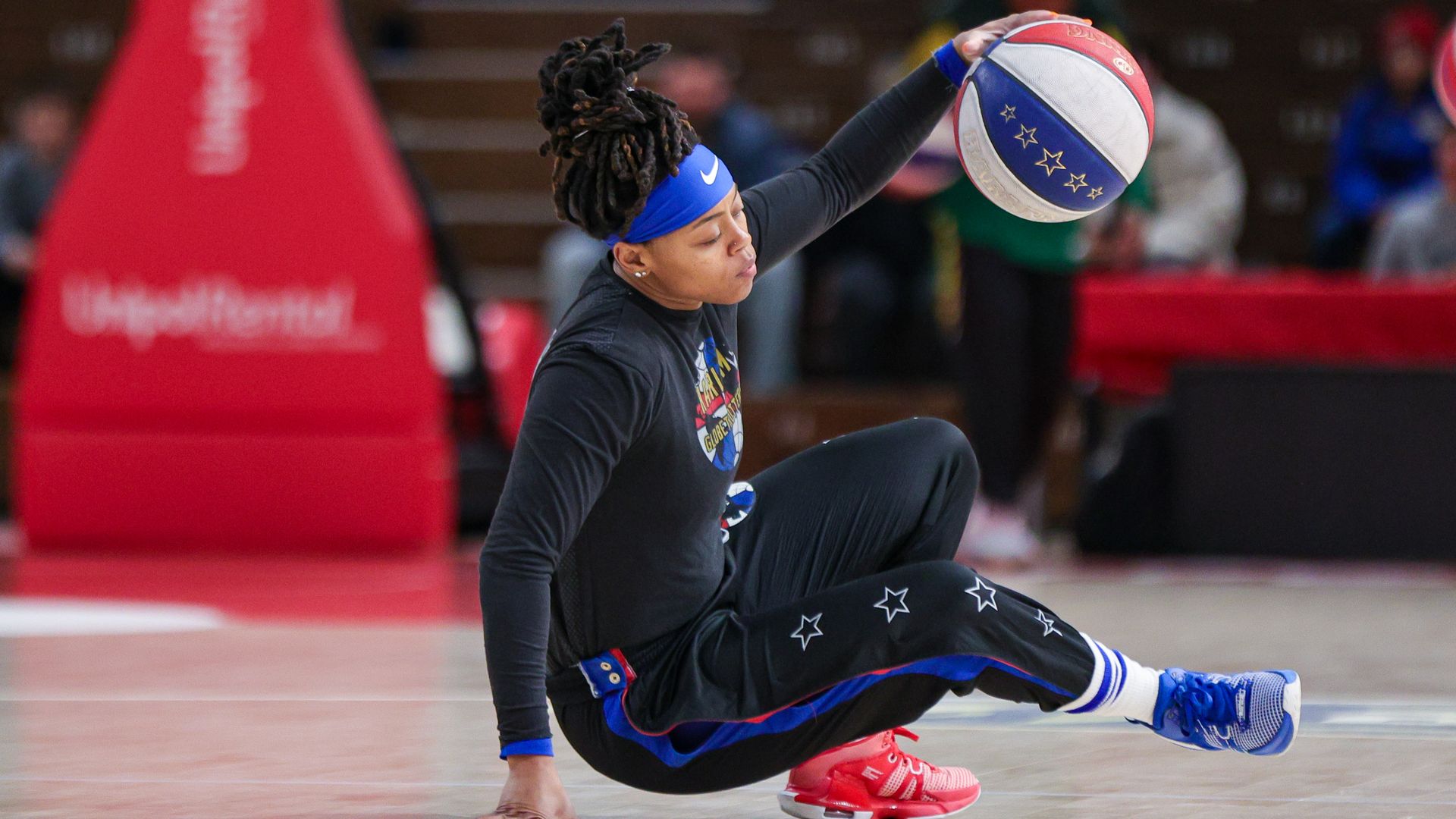 A woman uses one hand to dribble a red, white and blue basketball decorated with stars while she holds herself up from the ground using another hand. 