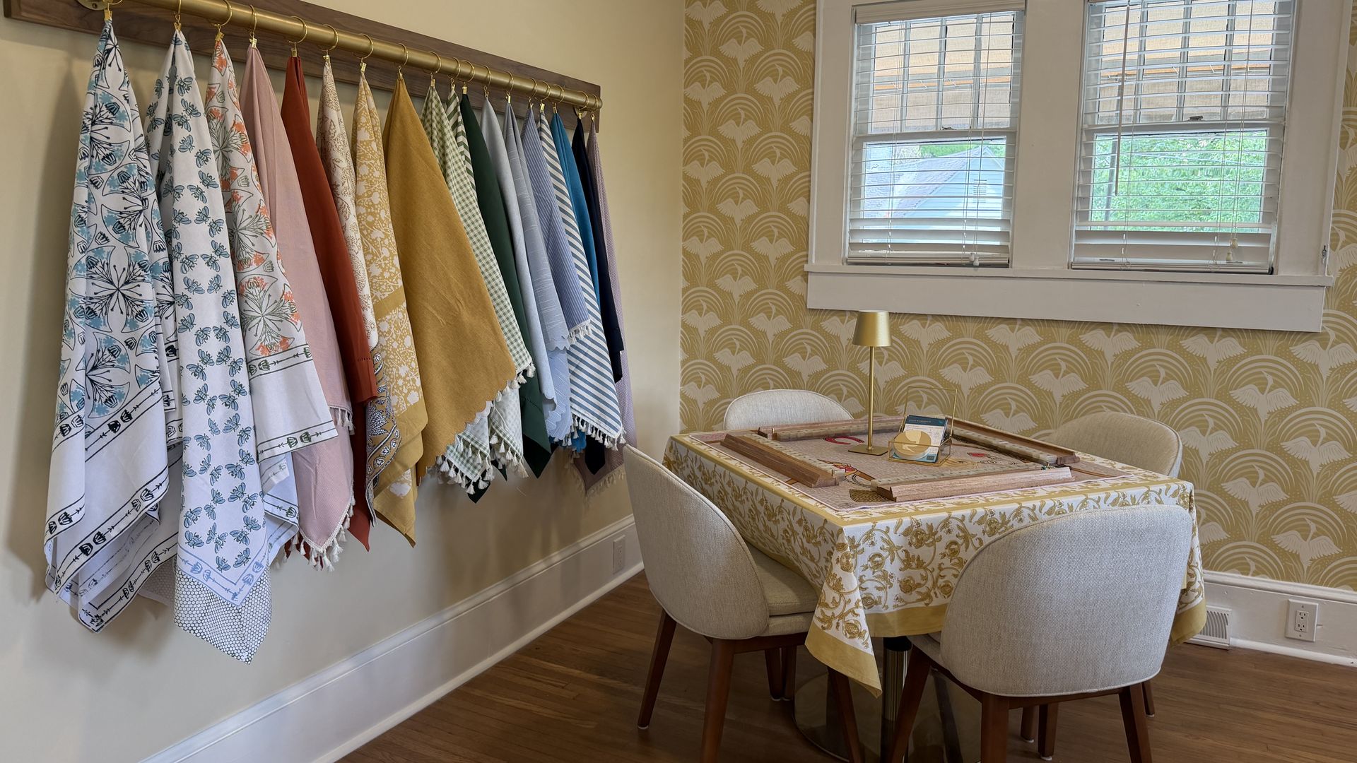 Cozy room with a brass towel rack holding colorful patterned towels along a beige wall; gold arched wallpaper, white blinds on a window, and a table with a yellow floral cloth.
