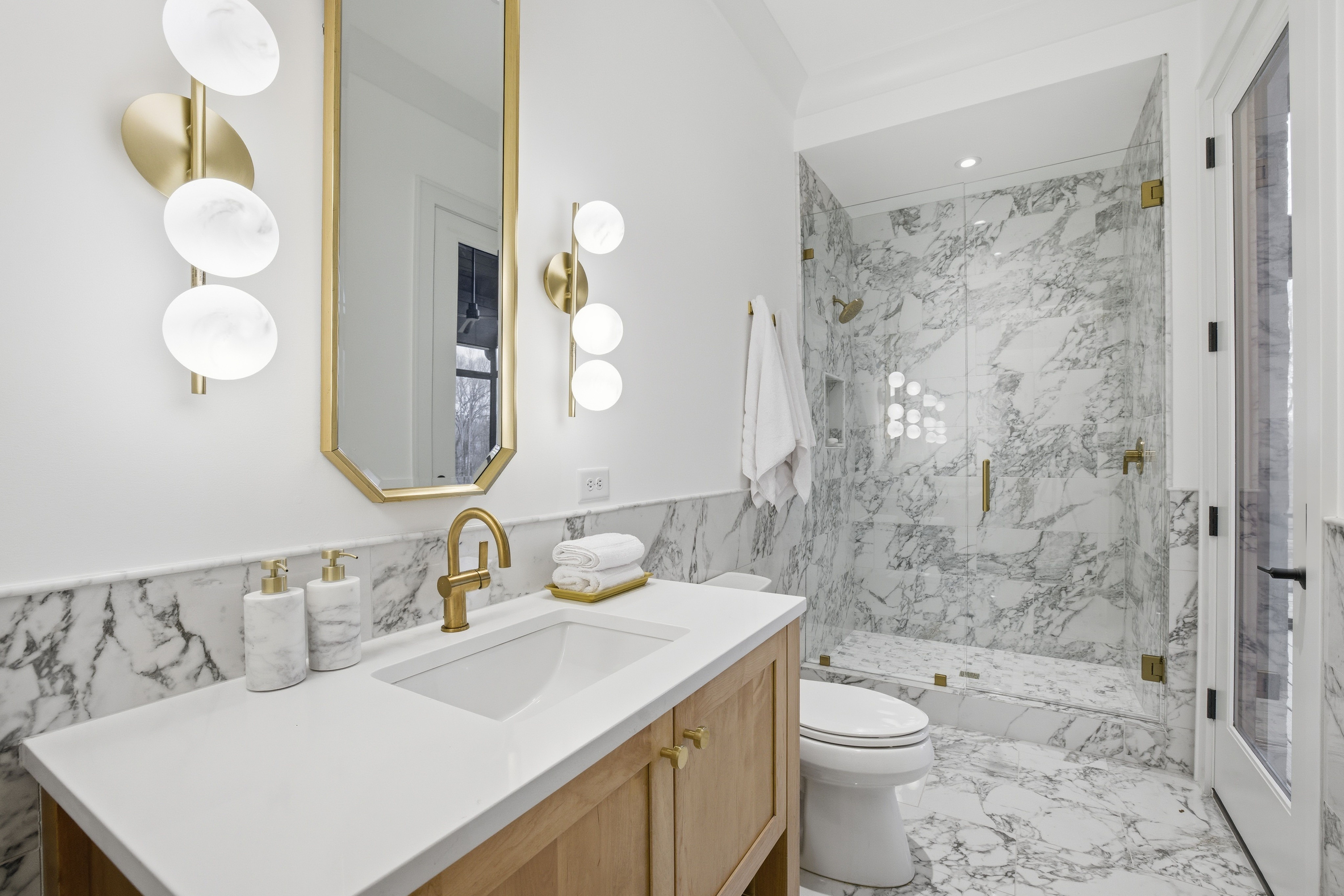 Modern bathroom with white marble tiles, gold fixtures, a glass shower, wood vanity with white countertop, two wall sconces, and a vertical gold-framed mirror.