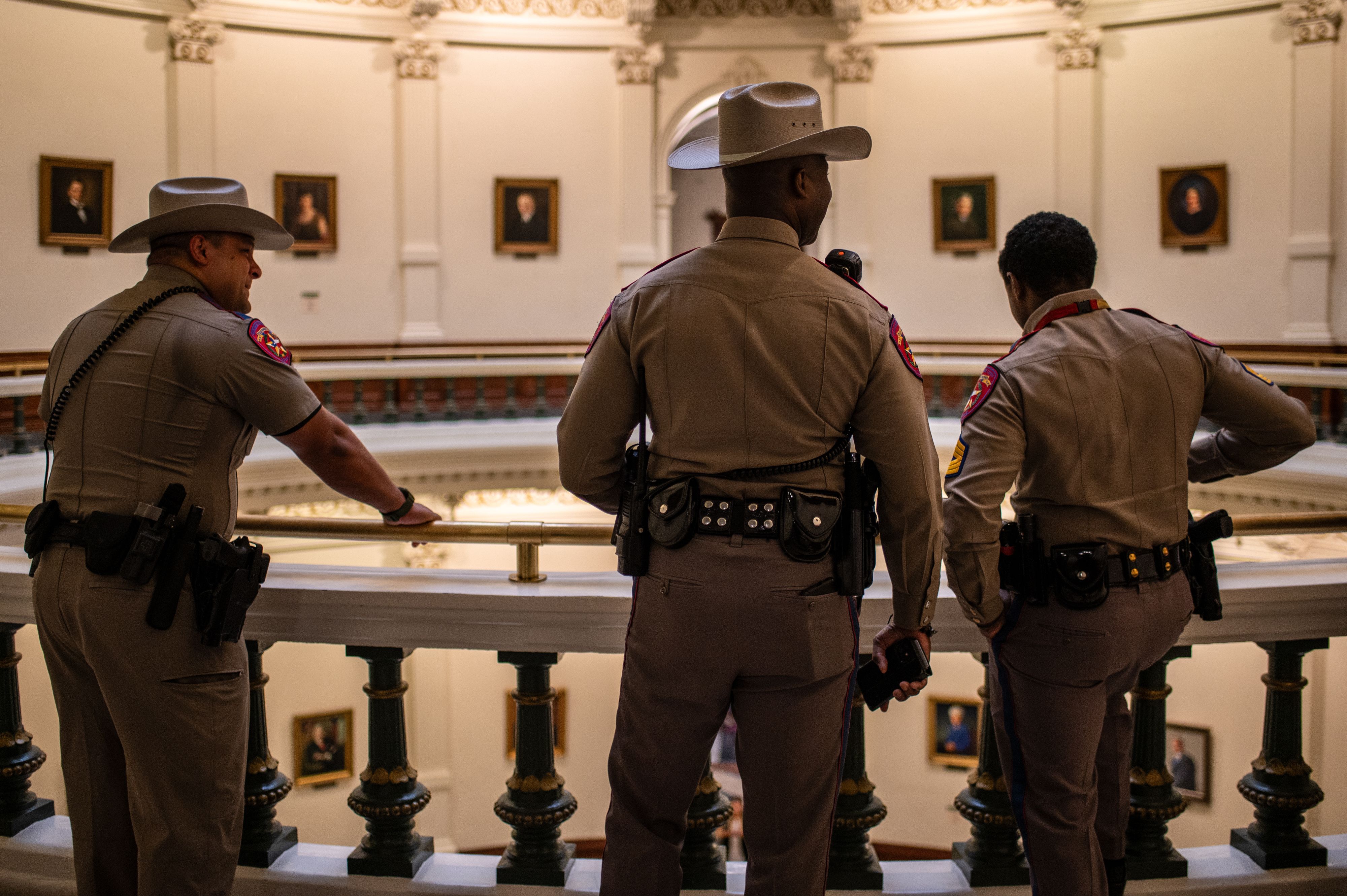 Three uniformed police officers, two wearing hats, stand facing away on an ornate balcony inside a historic building with framed portraits and decorative columns.