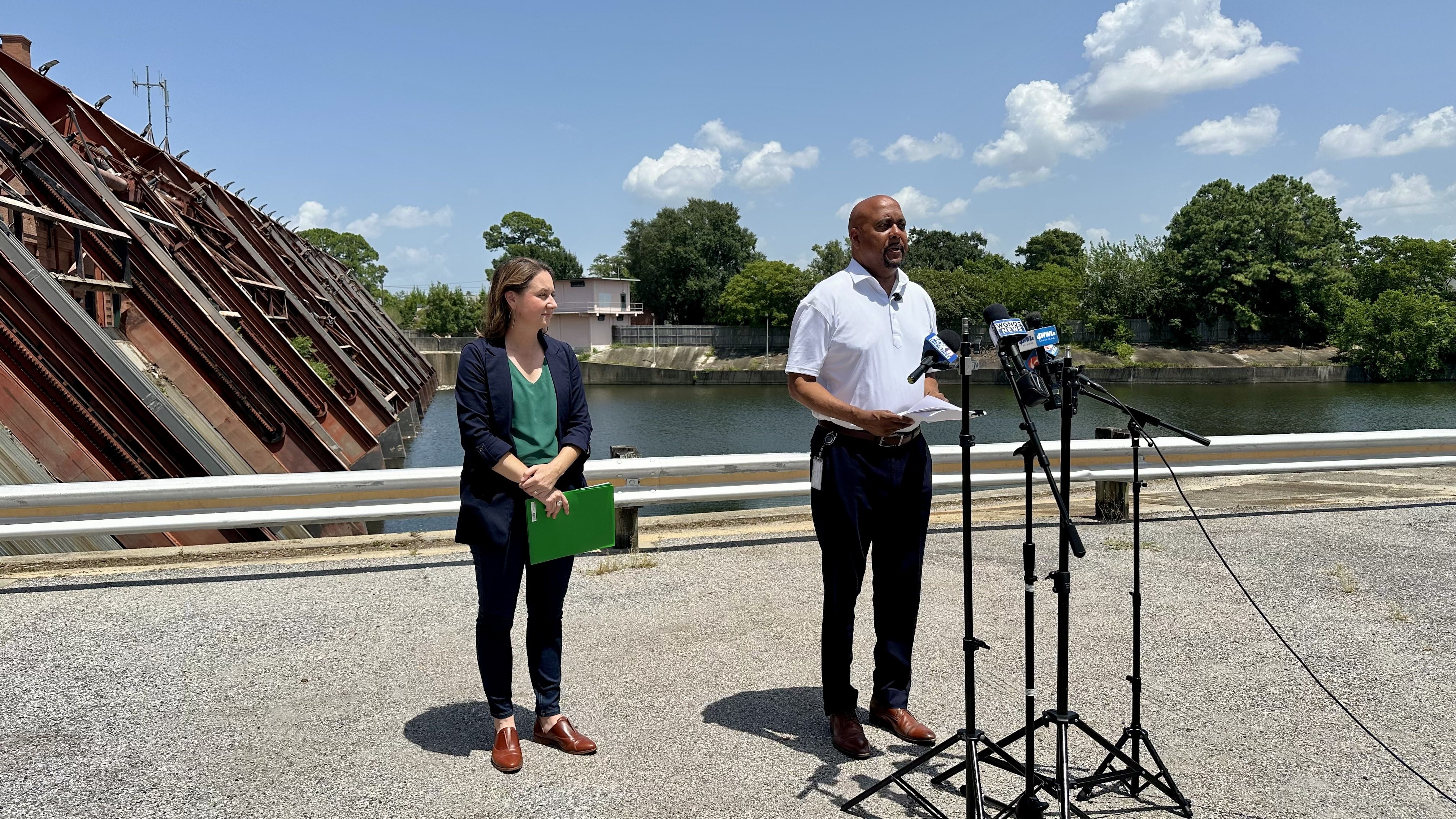 Man in a white polo speaks into multiple microphones while a woman with a green folder stands beside him by a river. A red dam slants on the left; trees and blue sky with clouds fill the background.