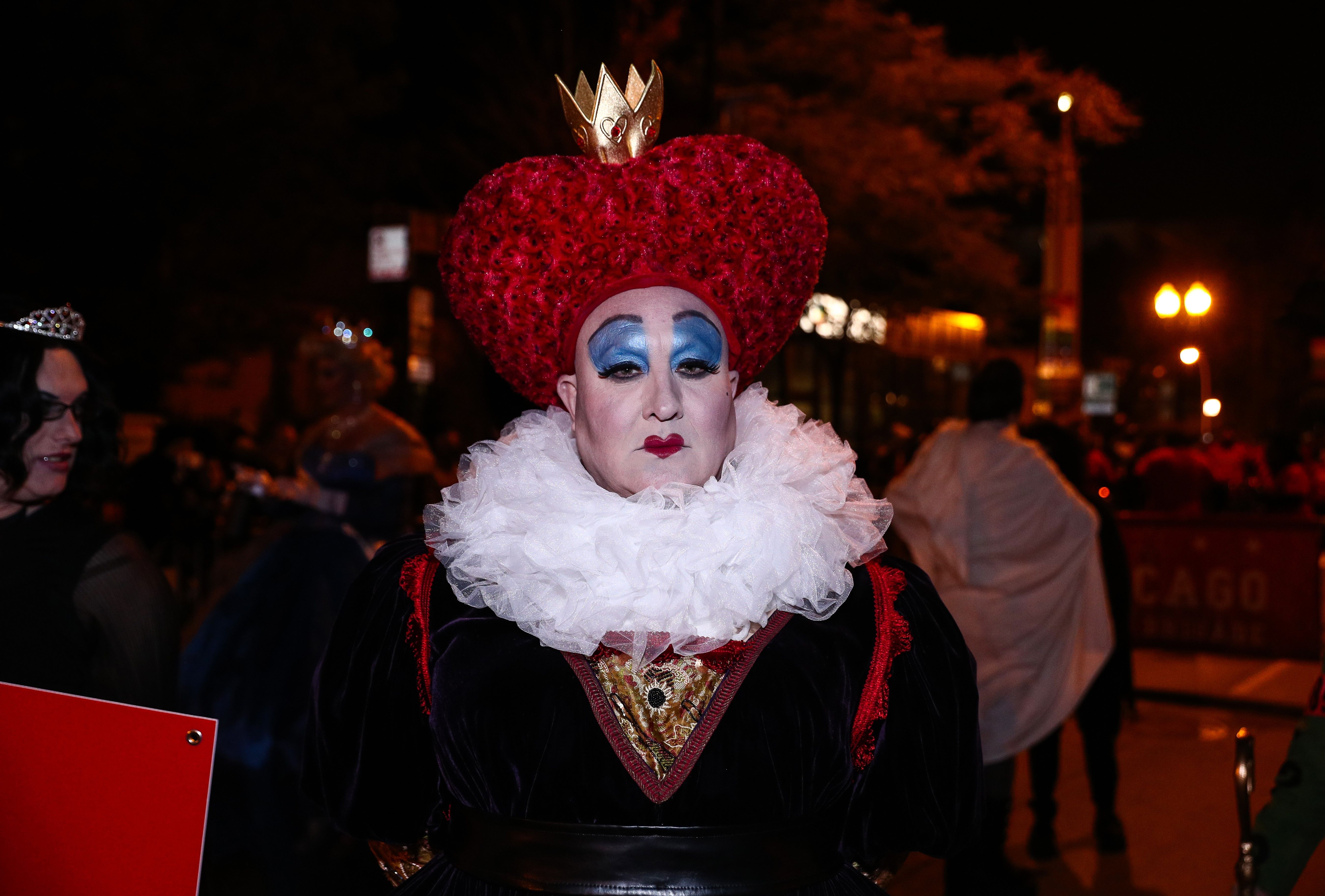 Person in dramatic makeup with blue eyeshadow and red lips, wearing a red heart-shaped headdress topped by a gold crown, white frilly collar and dark velvet costume; night street scene.