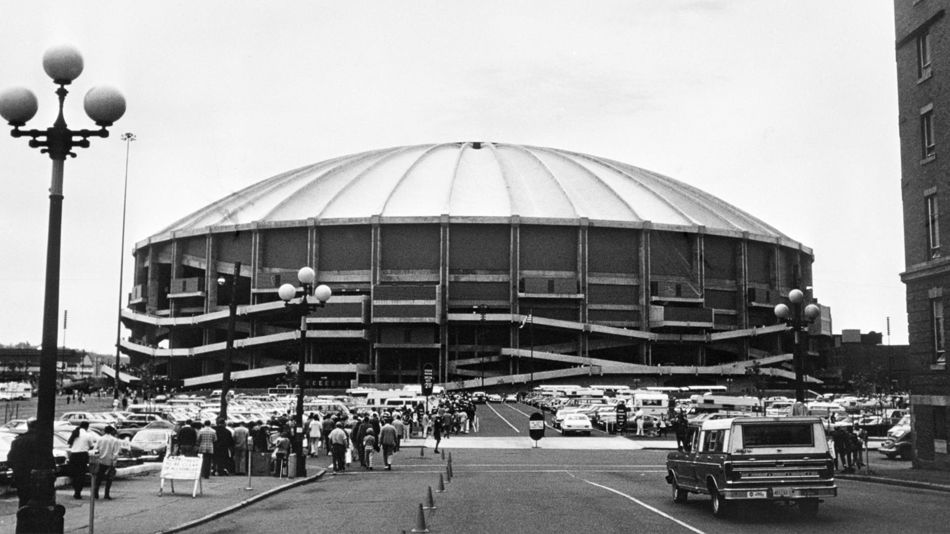 The Kingdome is shown in a black and white photo from when it was newly built, with people flocking toward the entrances.