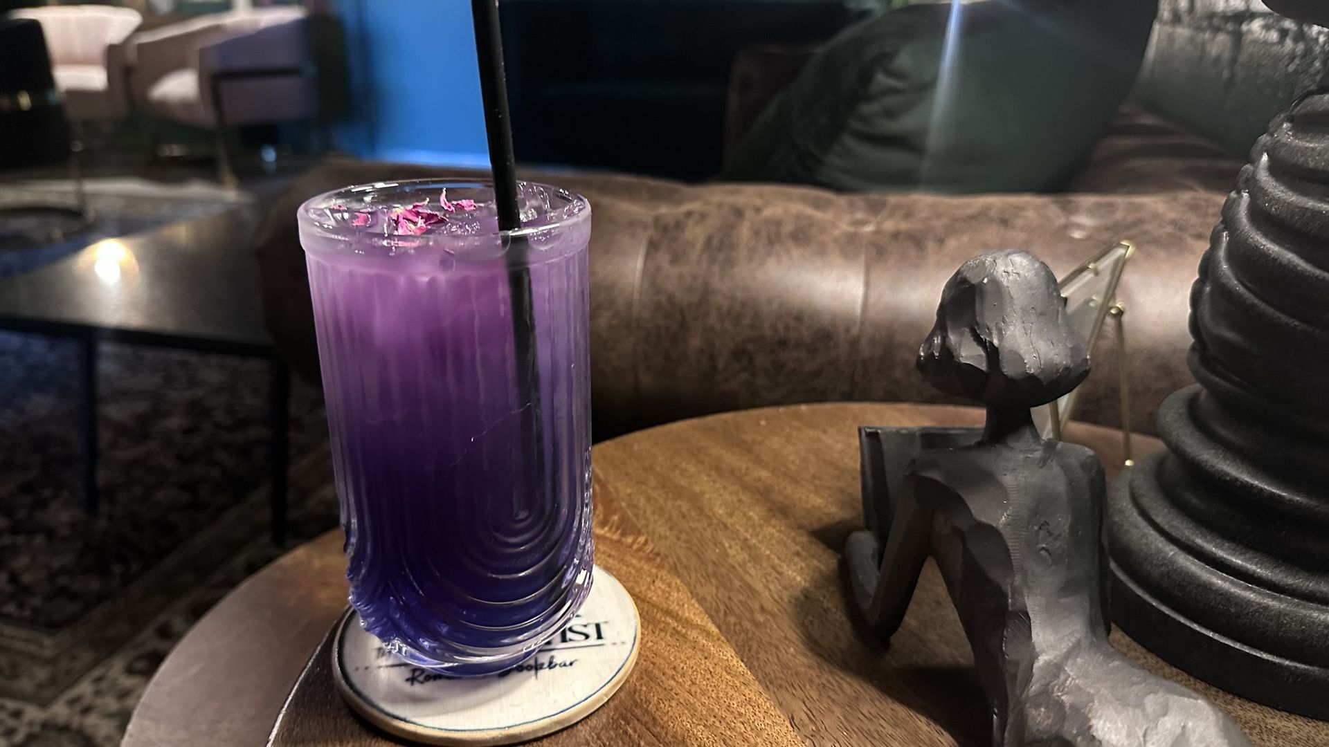 Purple layered iced drink with petals on top in a clear glass with black straw on a round wooden table next to a black abstract human sculpture and dimly lit lounge area in background.