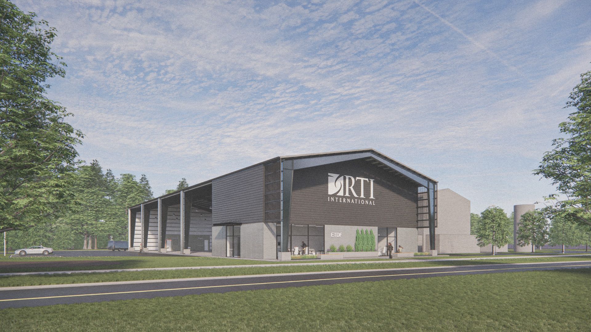 Large modern gray and black RTI International building with open bays, surrounded by green grass, trees, and a road in front under a blue sky with scattered clouds.