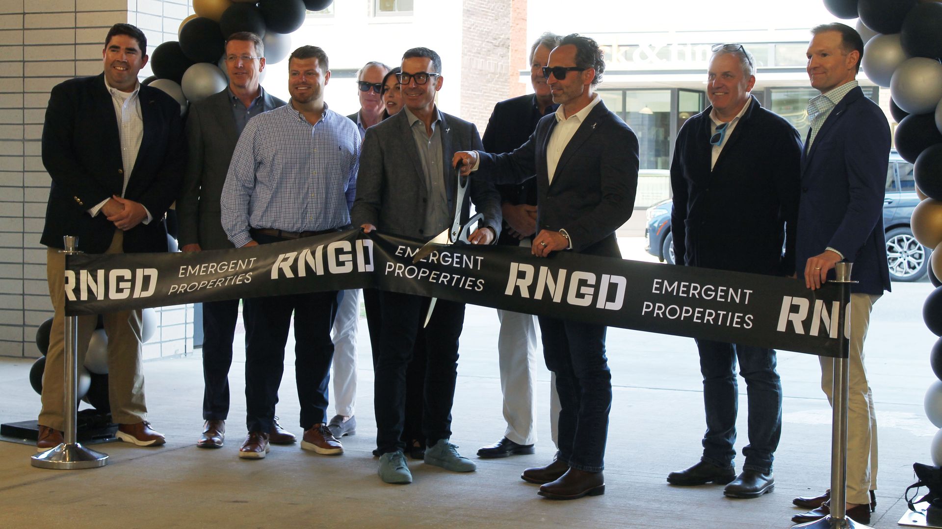 A group of men in suits cut a ribbon at a ceremony; the black ribbon reads "RNGD EMERGENT PROPERTIES" as a black-and-gold balloon arch frames the scene.