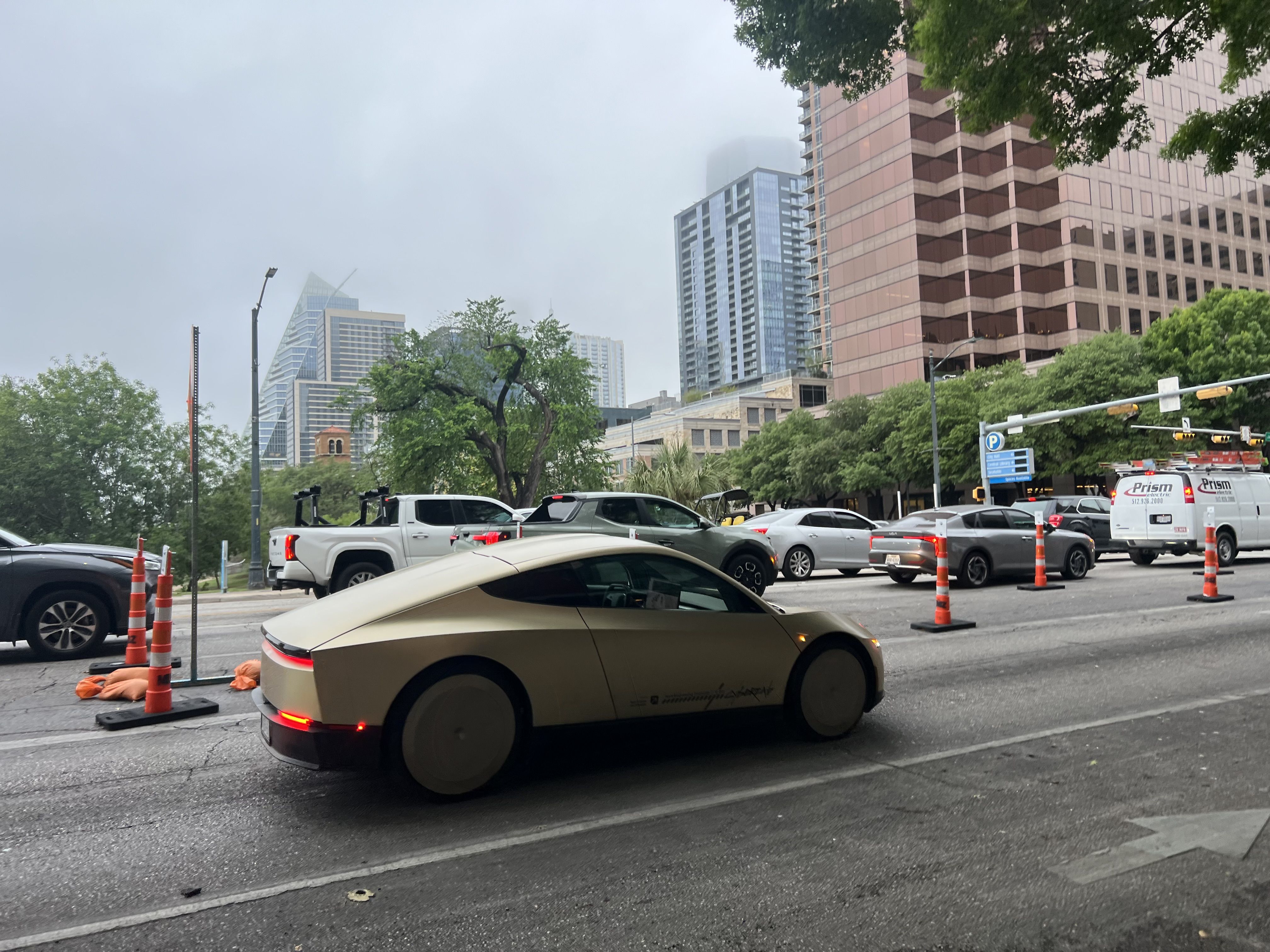 A sleek beige futuristic sedan drives along a city street lined with orange construction cones. In the background are modern glass skyscrapers, green trees, and an overcast sky.
