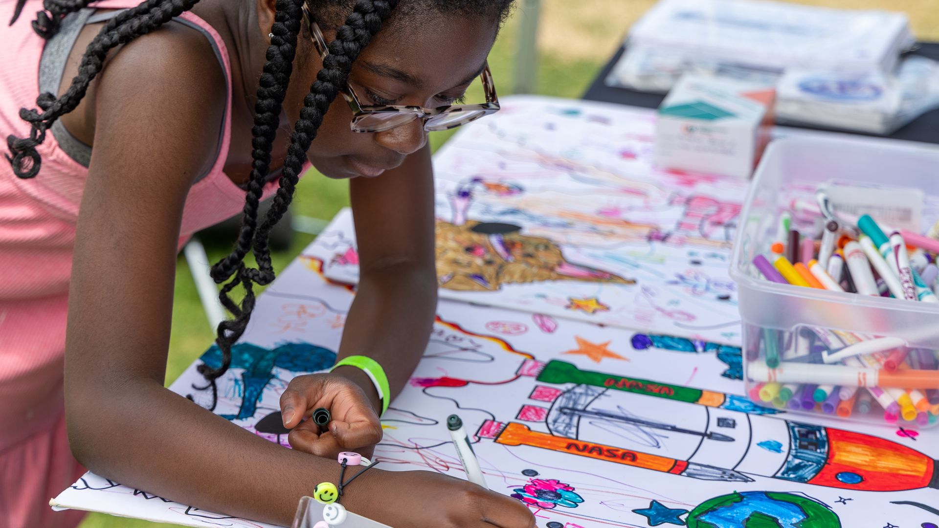 A young girl colors in a picture of a rocket on a table with a box of markers nearby. 