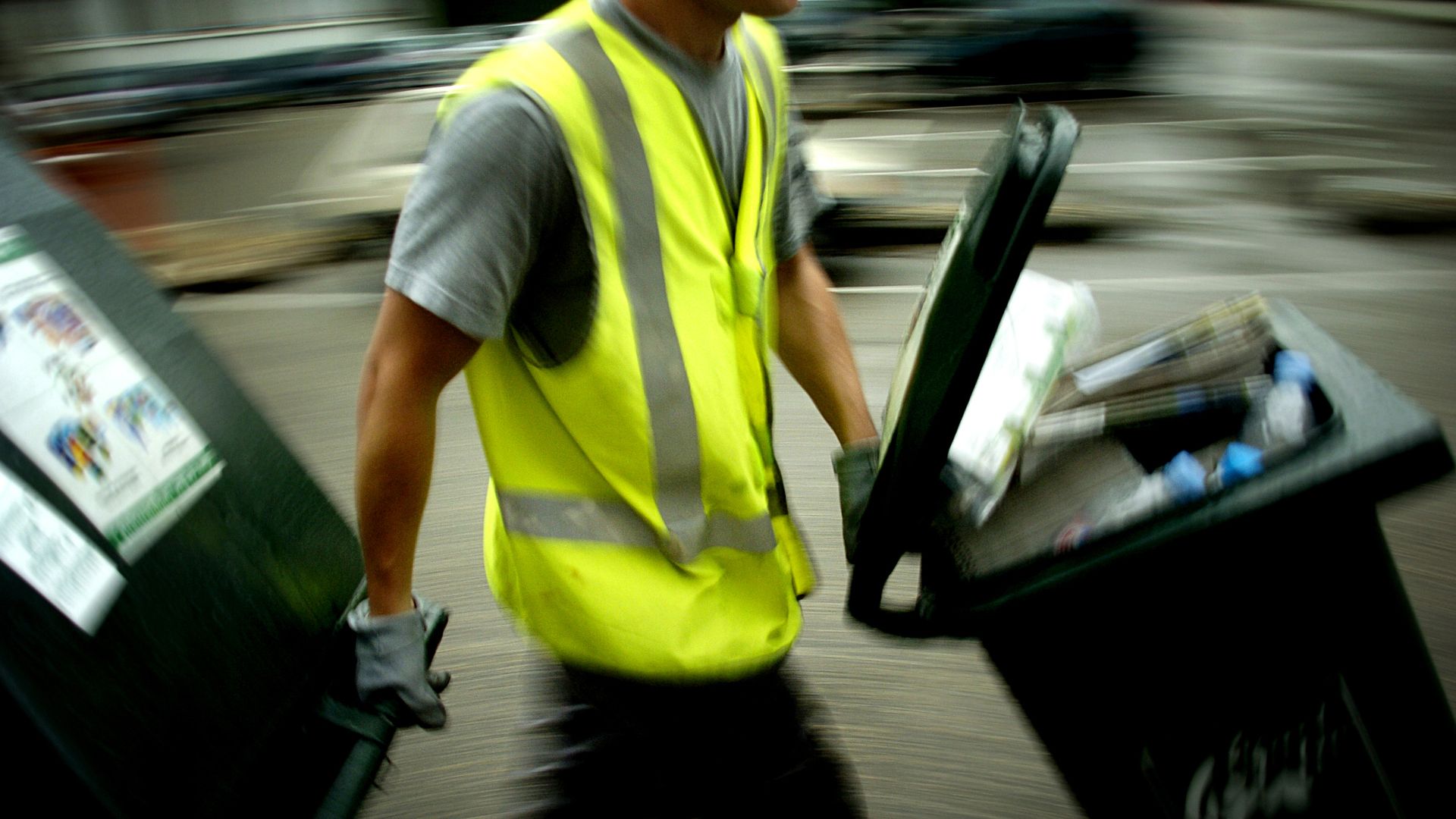 A man in a yellow vest moves two large plastic bins of rubbish.