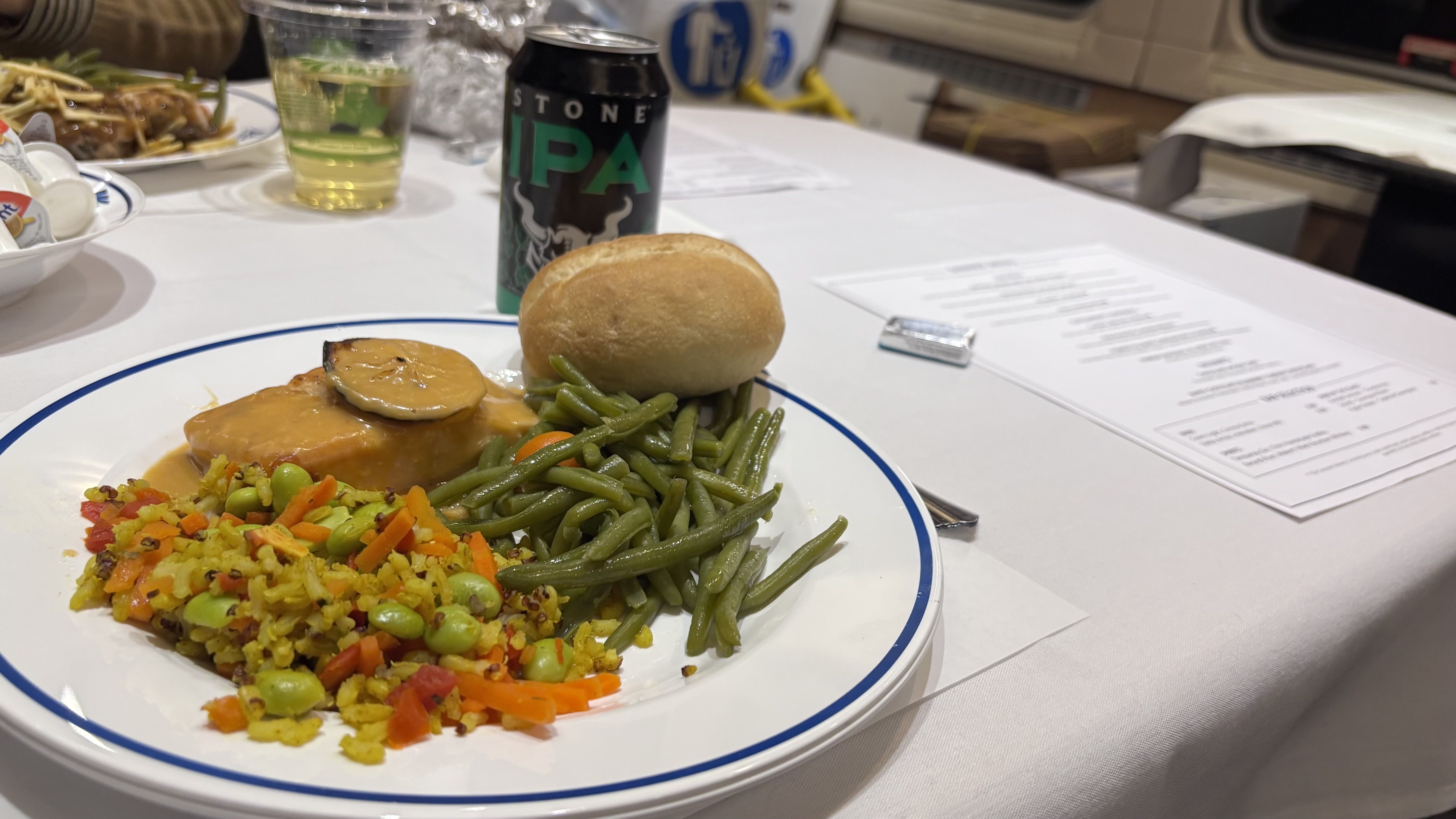 Plate with green beans, yellow rice with vegetables, a bread roll, and meat with sauce and a lemon slice. Stone IPA beer can and a drink cup on a white table with papers.