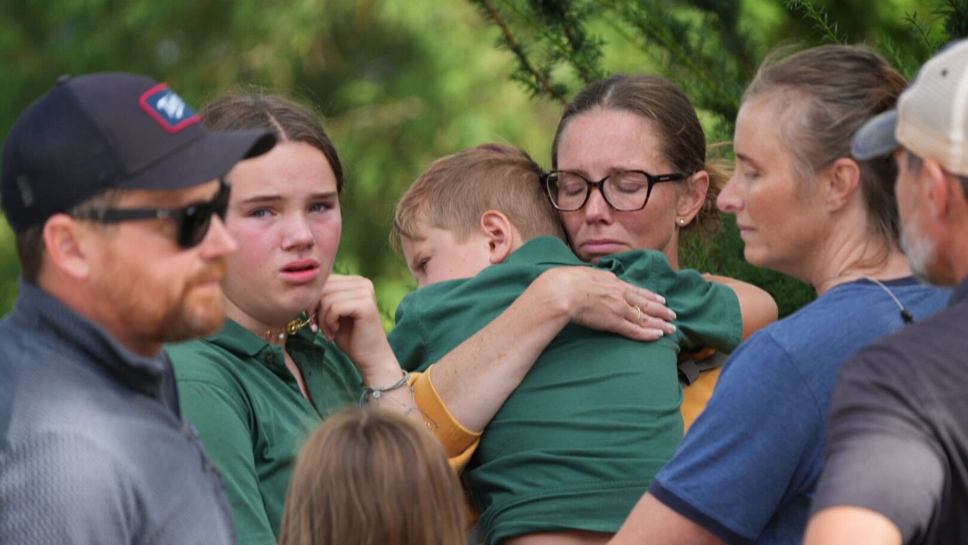 A group of people outdoors, including a woman with glasses hugging a young boy wearing green, a tearful girl in green, and others showing concerned and sad expressions.