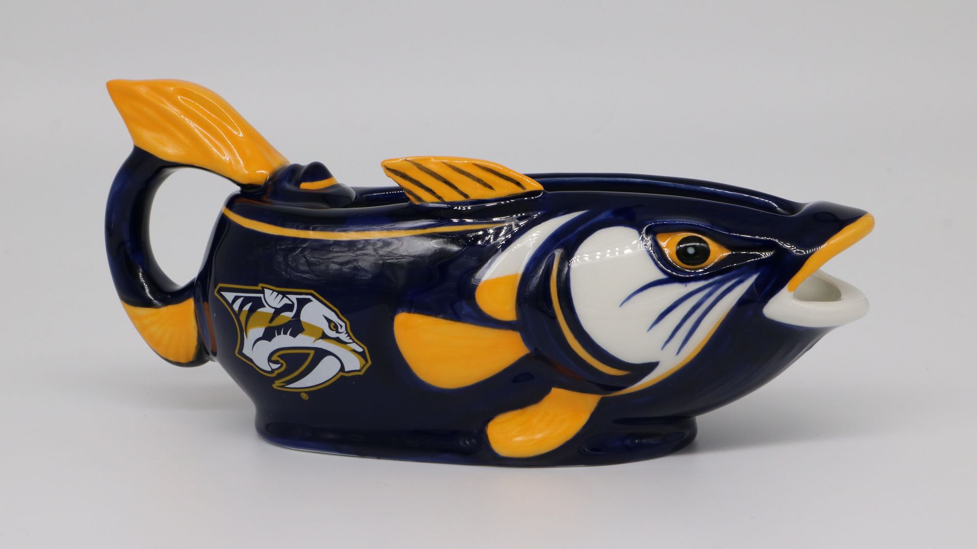 Glossy blue fish-shaped ceramic mug with orange fins and yellow accents, a white mouth, and a yellow eye; side features a yellow emblem. Handle on the left against a plain light background.