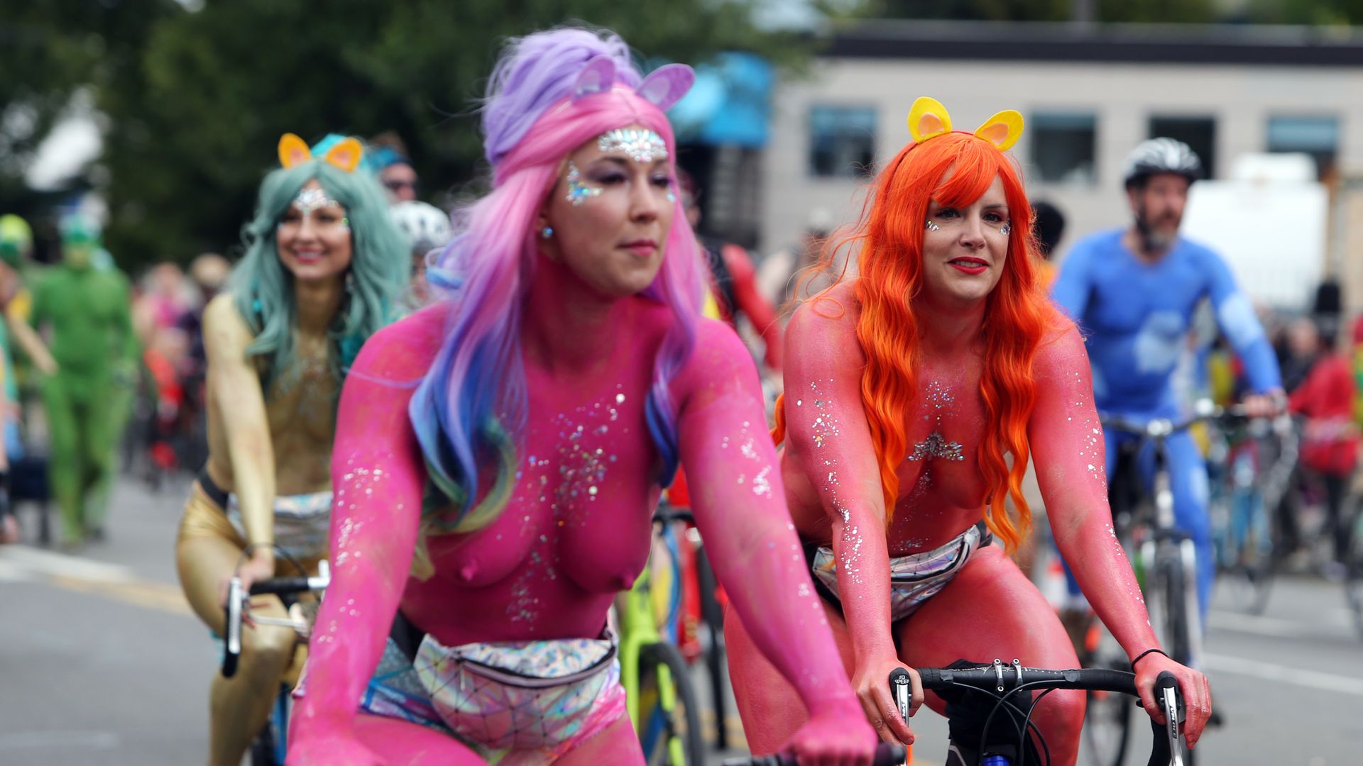 Topless women covered in bright body paint, sparkles and cat ears ride bicycles in a parade.
