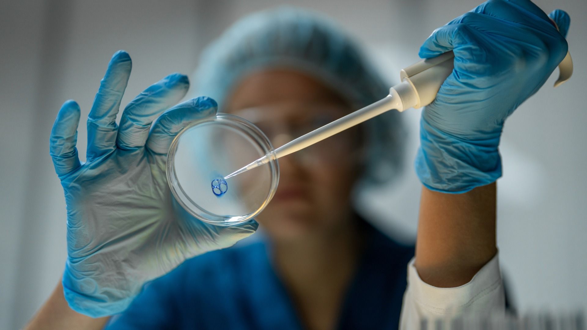 Close-up of a lab technician wearing blue gloves and a hairnet, using a pipette to transfer liquid into a Petri dish with a blue circular specimen, in a sterile lab setting.