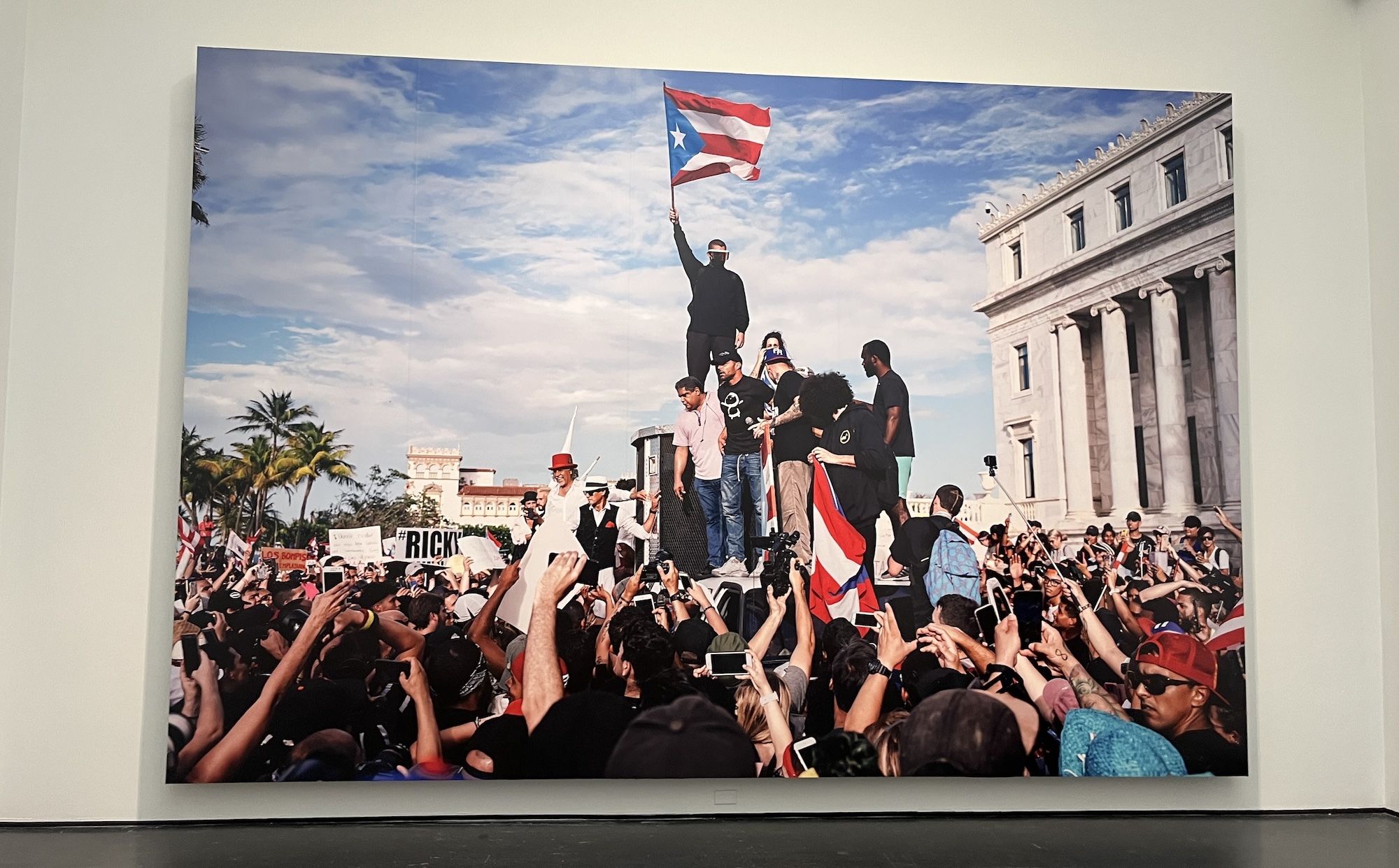 Massive crowd of protesters surrounds a raised platform where a man waves a Puerto Rican flag; onlookers film with phones as a columned building looms nearby, under a blue sky.
