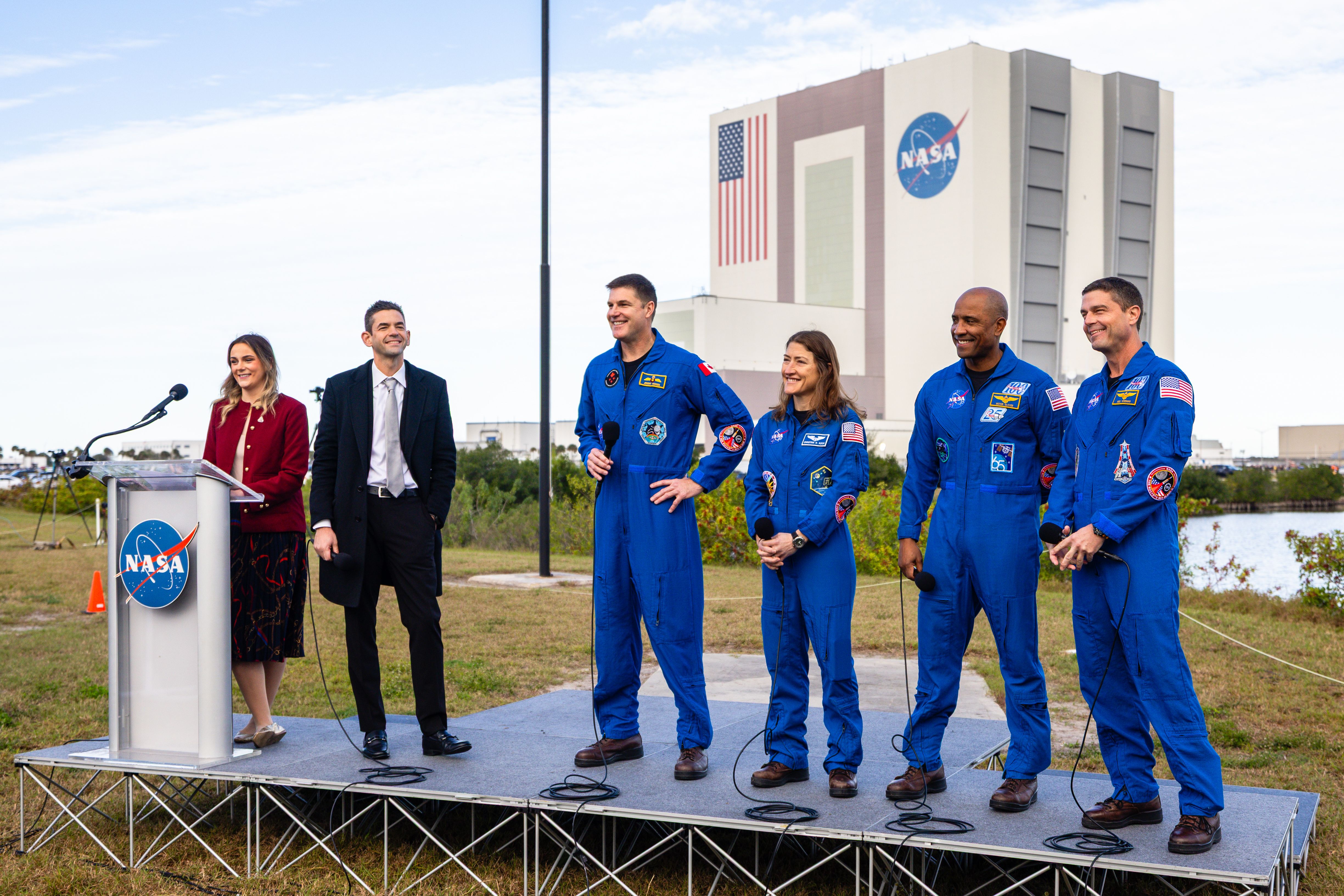 Image shows astronauts, dressed in blue flight suits, holding microphones and talking on a stage.