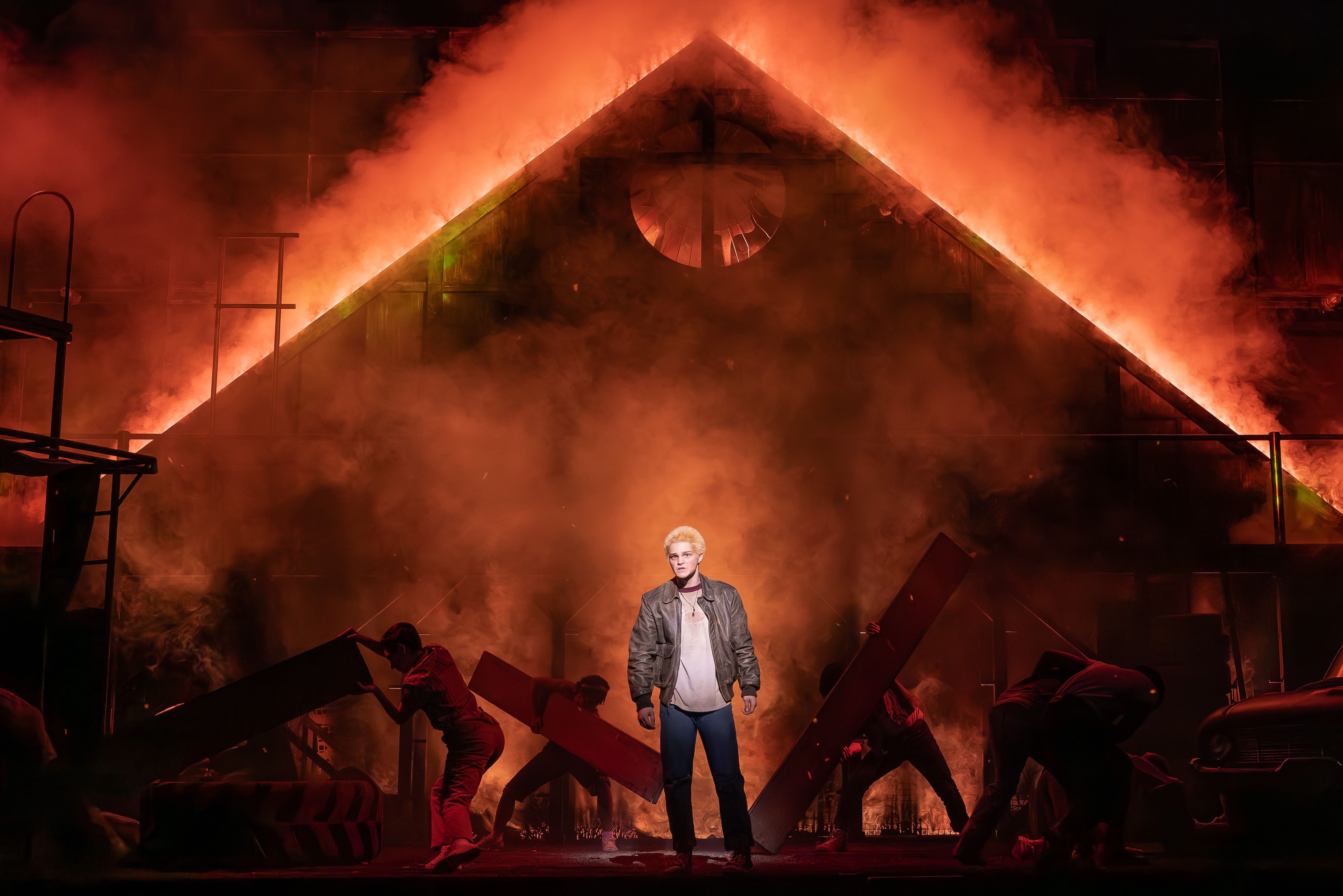 A nighttime stage scene with intense orange flames and smoke engulfing a triangular theater set. A person in a leather jacket stands center foreground as dancers move in the smoky background.