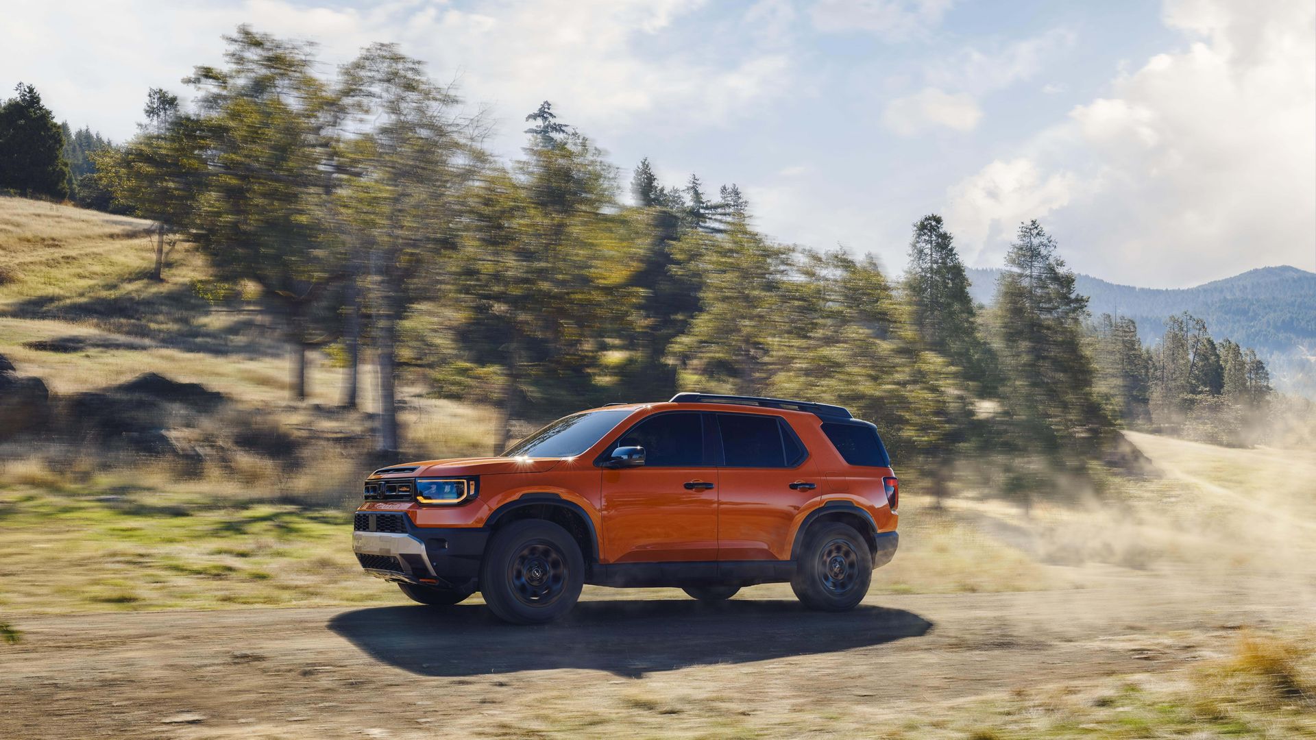 Orange Honda Passport SUV driving fast on a dirt road in a green, hilly landscape.