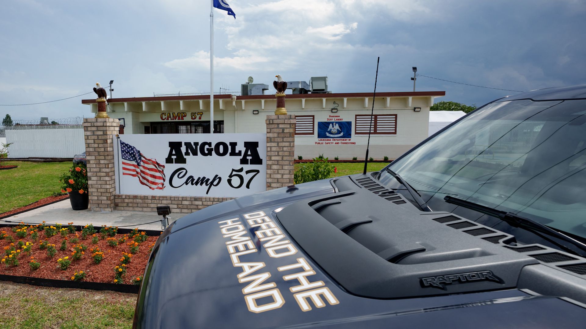 A sign for "Angola Camp 57" sits in front of a building with the same title. A car is seen in the foreground with "Defend the homeland" printed across the hood.
